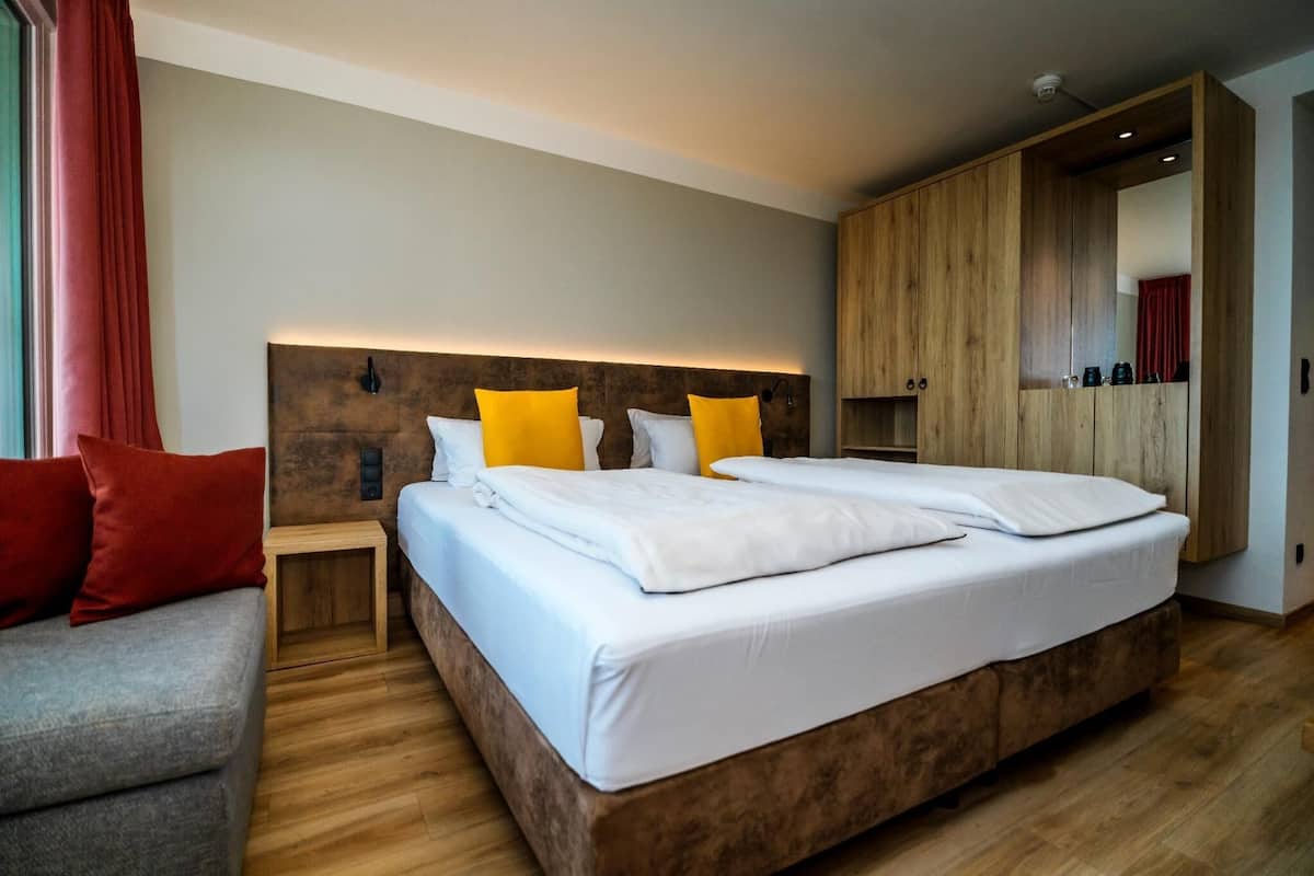 Una habitación de hotel moderna con una cama grande, un cabecero de madera, una mesita de noche pequeña y un sofá gris.