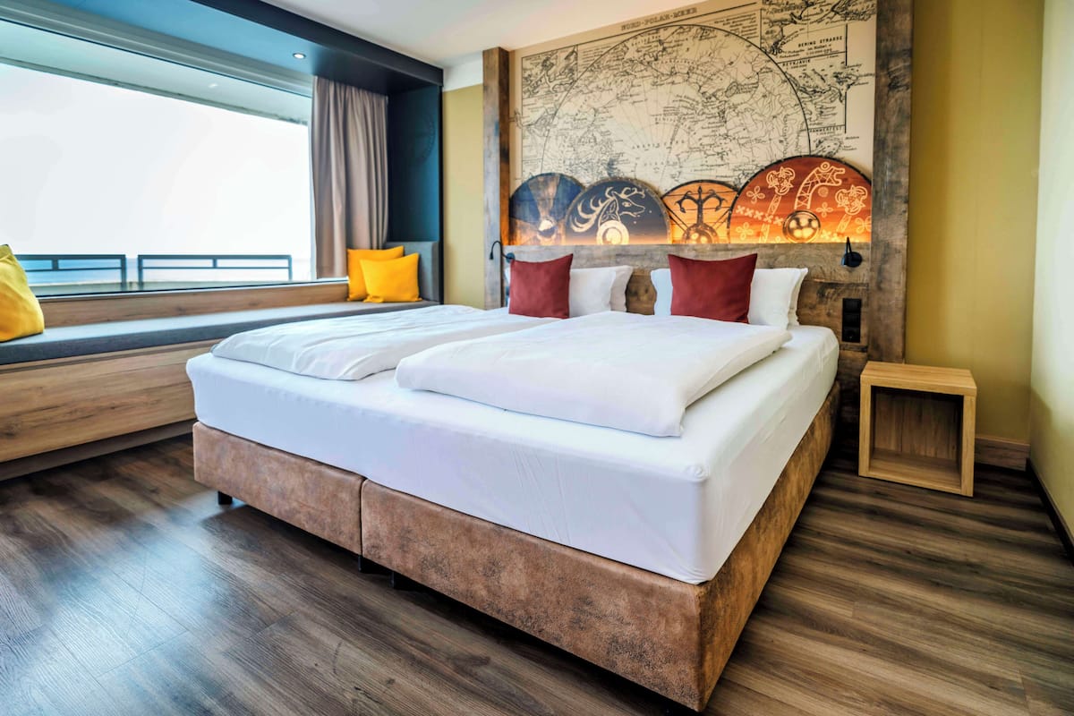 Un dormitorio moderno con una cama grande, un rincón con asiento junto a la ventana, un mural de un mapa y una pequeña mesita de noche de madera.