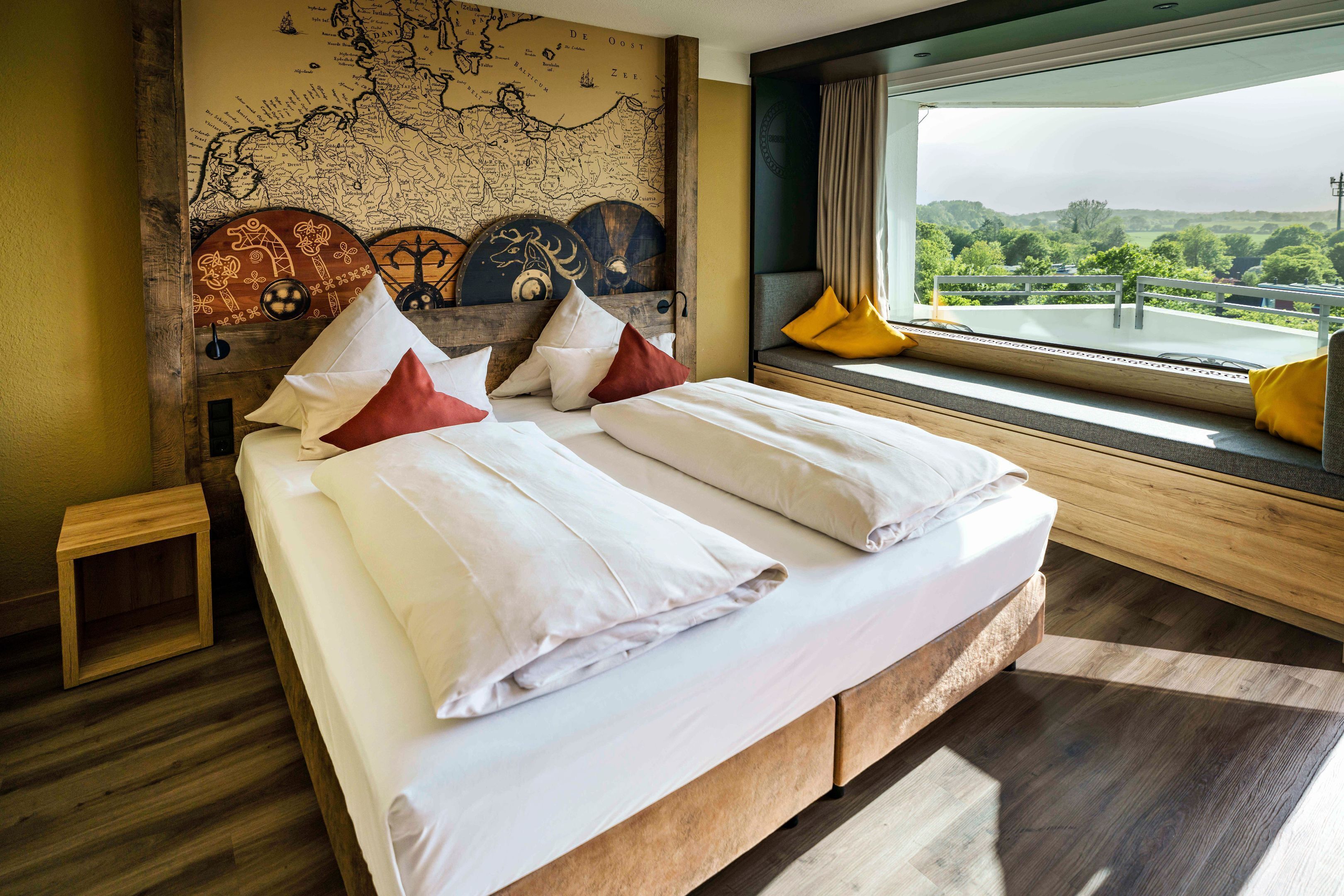 A double bed with white linens and red and white pillows, a wooden headboard with a map, a small wooden nightstand, and a window with a view of greenery.