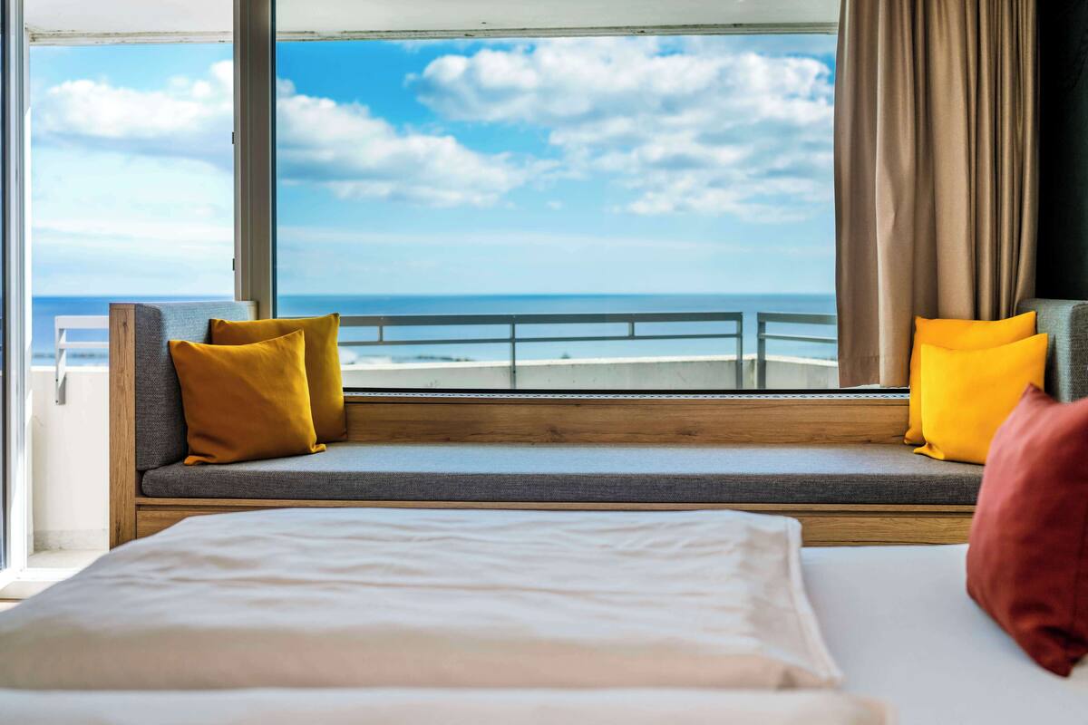 Un balcón con vistas al mar, una cama con almohadas y una ventana con cortinas.