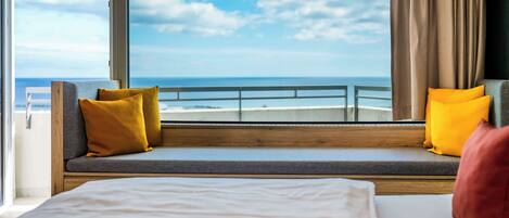 Een balkon met uitzicht op zee, een bed met kussens, en een raam met gordijnen.