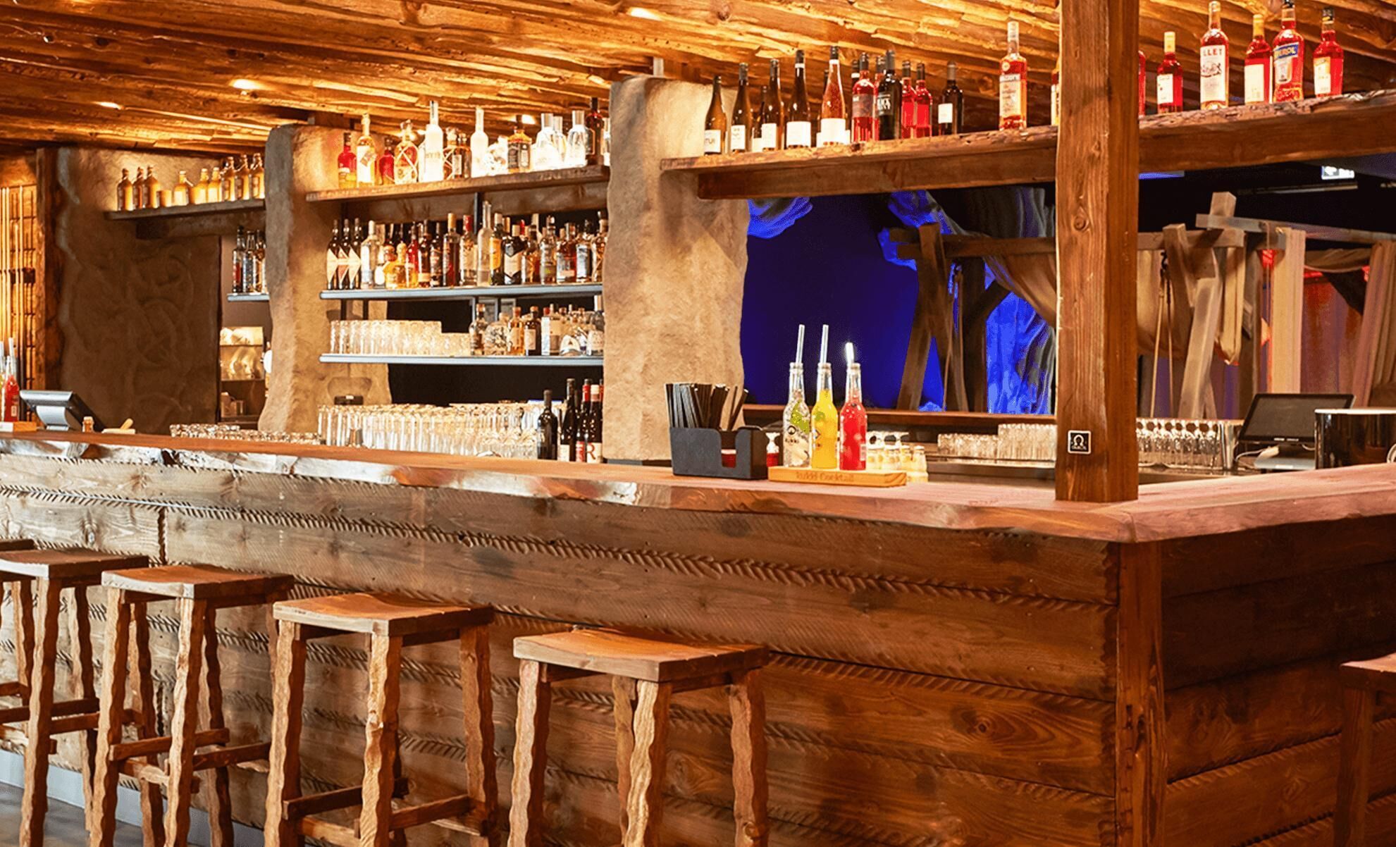 A bar area with wooden shelves stocked with various bottles, a bar counter, and wooden stools.