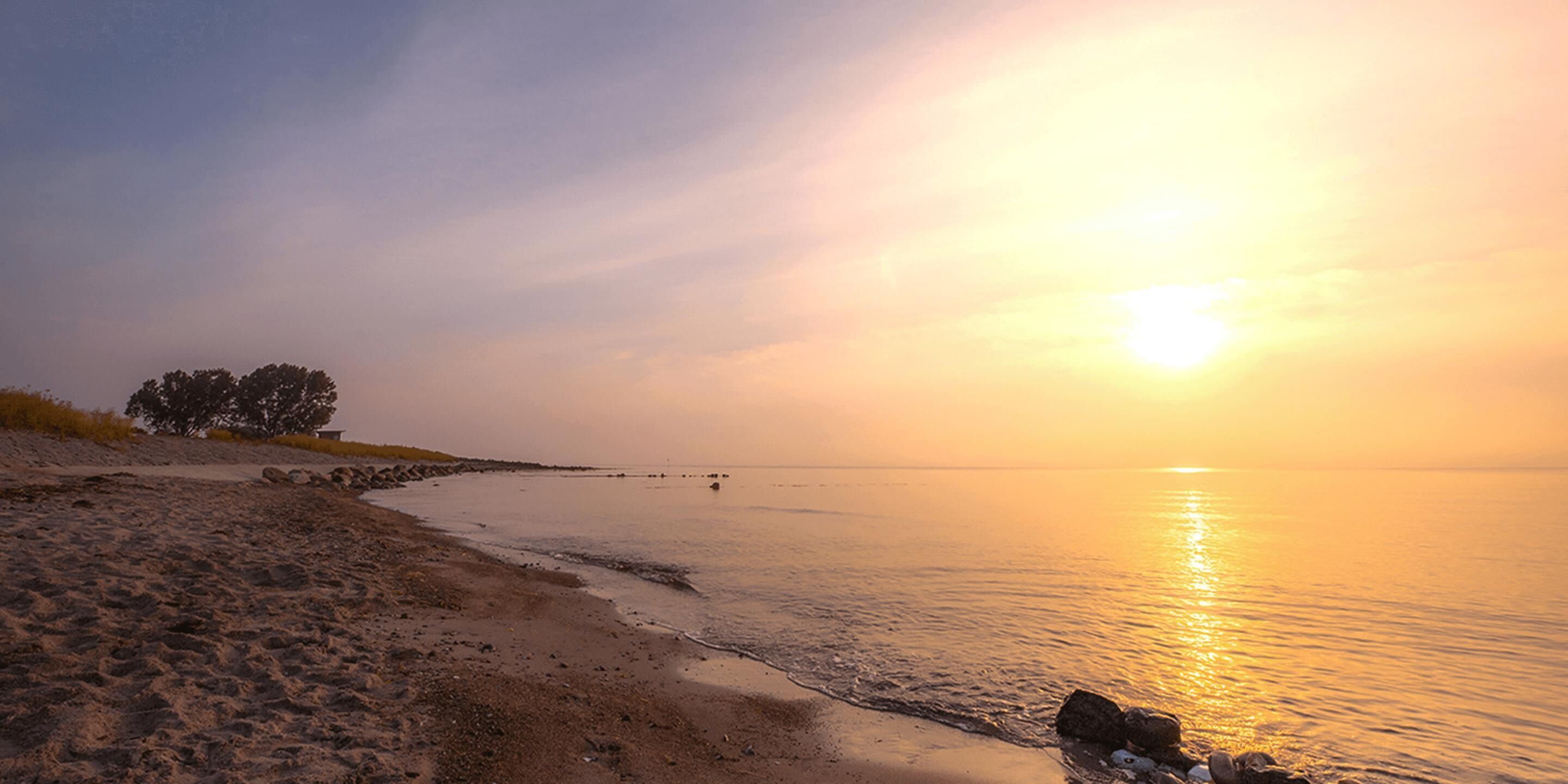 A beach with a sunset, calm water, and a few scattered rocks.