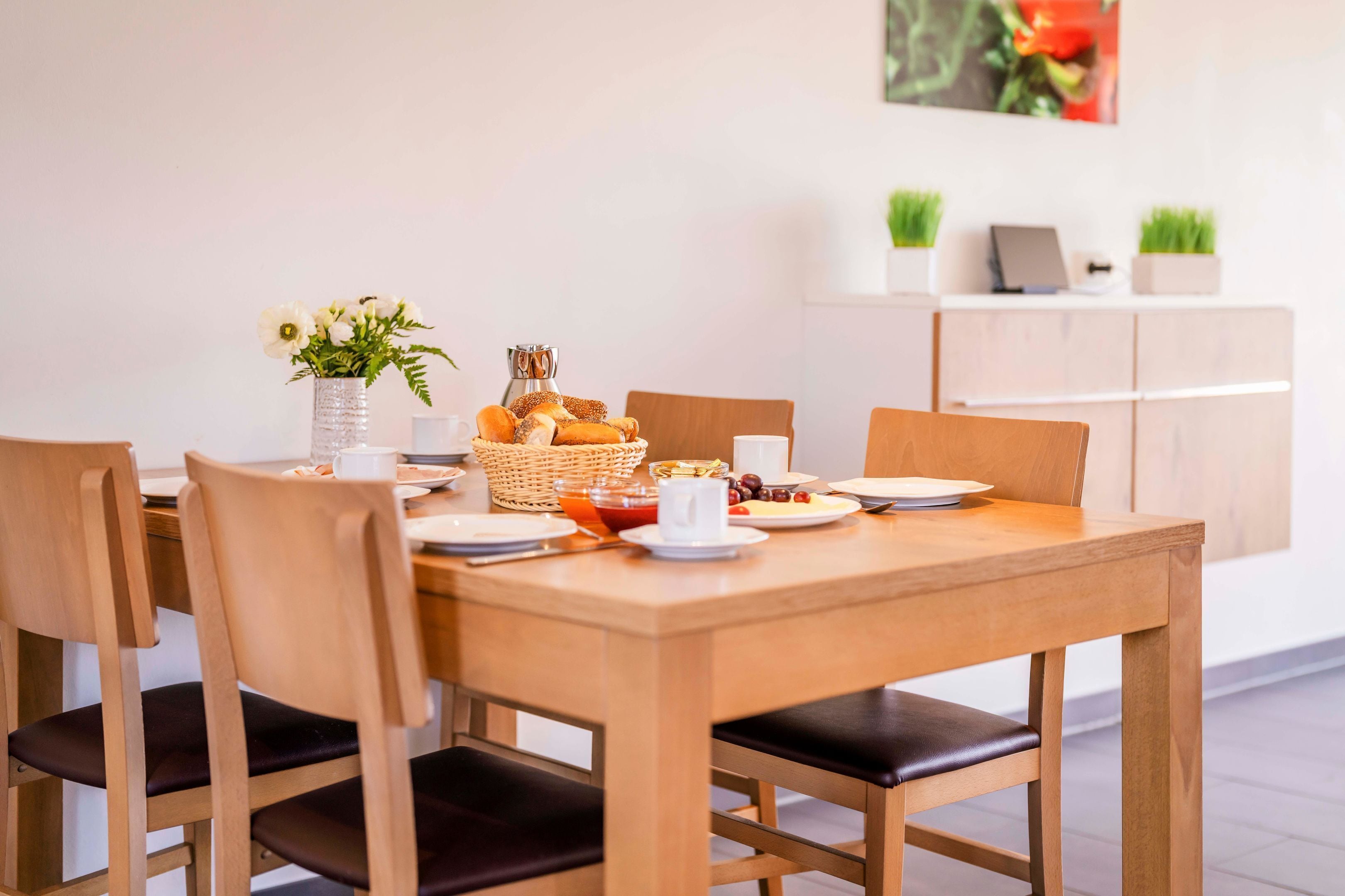 Una mesa de madera para comer con cuatro sillas, un jarrón de flores, una cesta de pan y una taza de café.