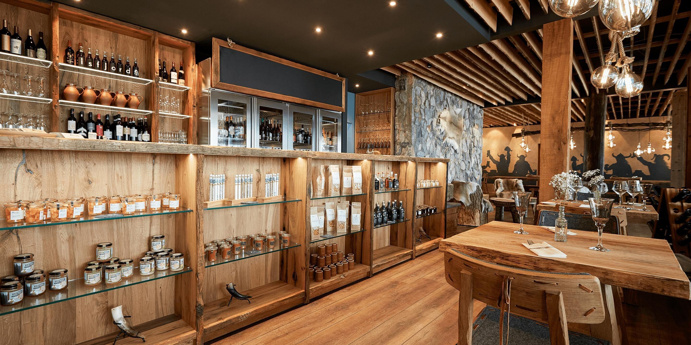 Interior de madera con estantes mostrando botellas y frascos, una mesa de madera con sillas y un mural en la pared.
