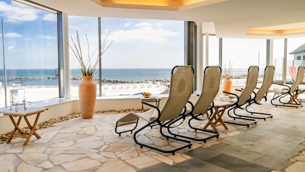 Una stanza luminosa con pavimento in pietra, ampie finestre vista spiaggia e diversi divani dove rilassarsi.