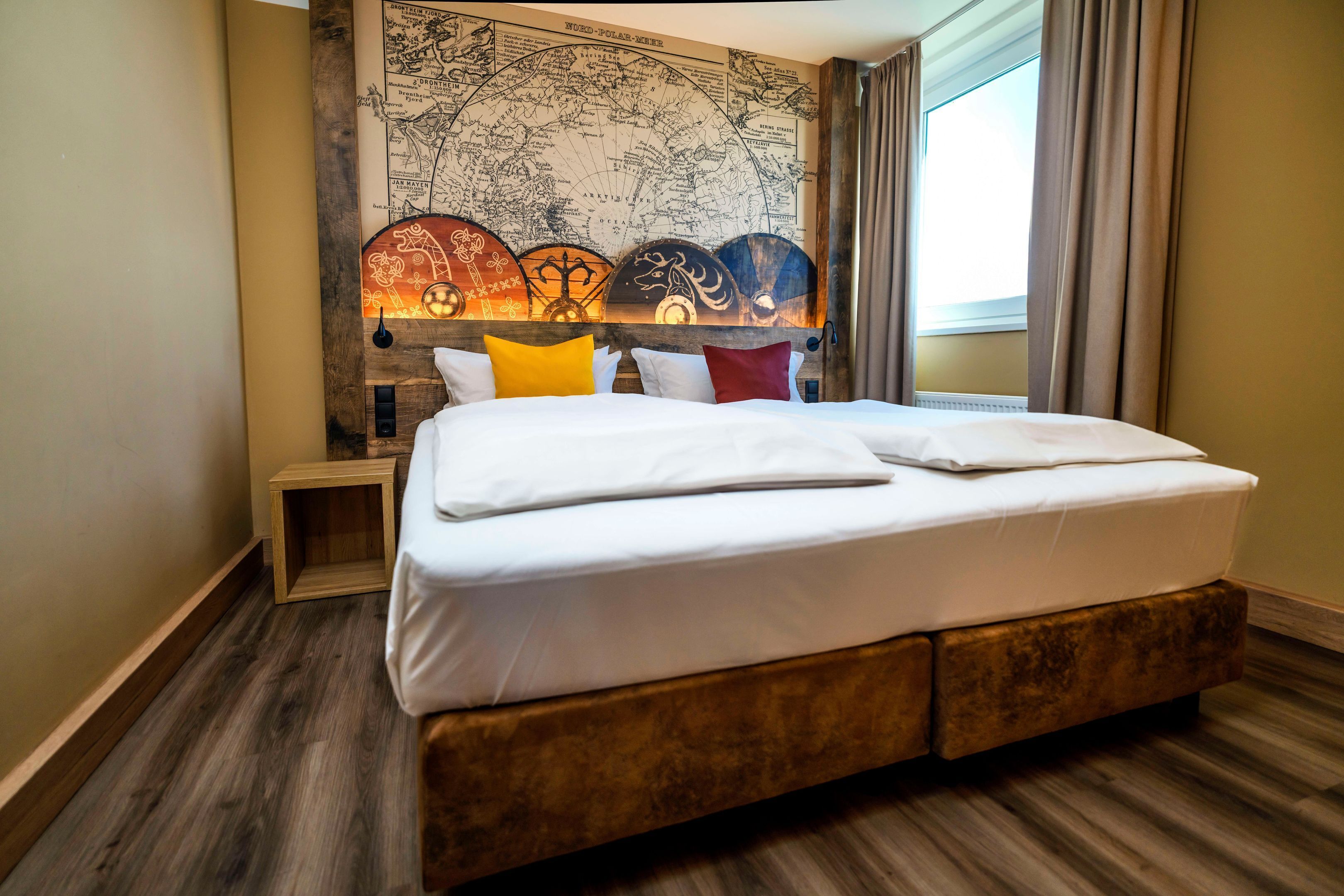Uma cama de casal bem arrumada com roupa de cama branca, cabeceira de madeira com luminária decorativa e um mural de mapa mundi na parede.