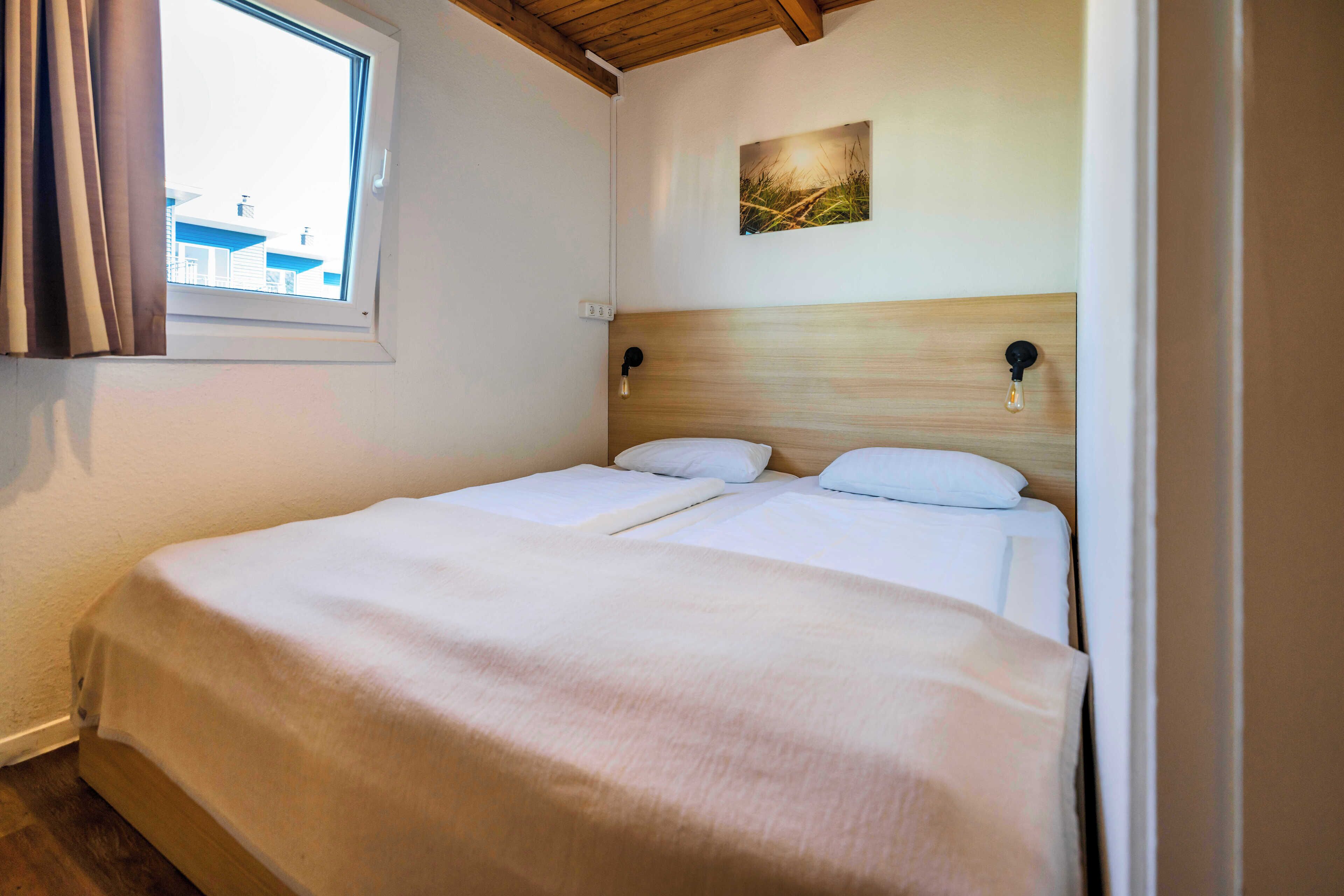 A double bed with a wooden headboard, a window with curtains, and a wall-mounted light fixture.
