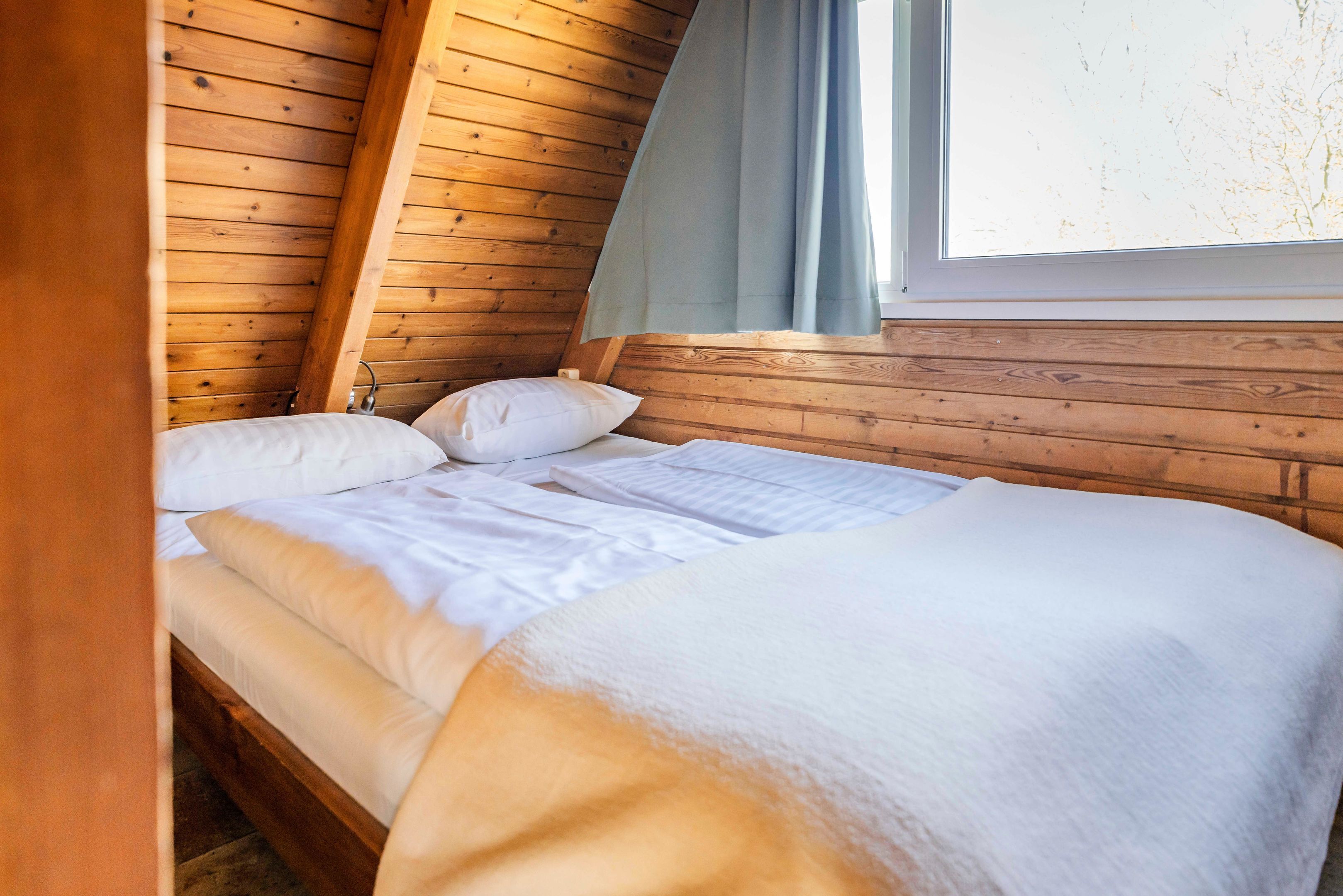 A neatly made bed in a wooden cabin room with a window and curtains.