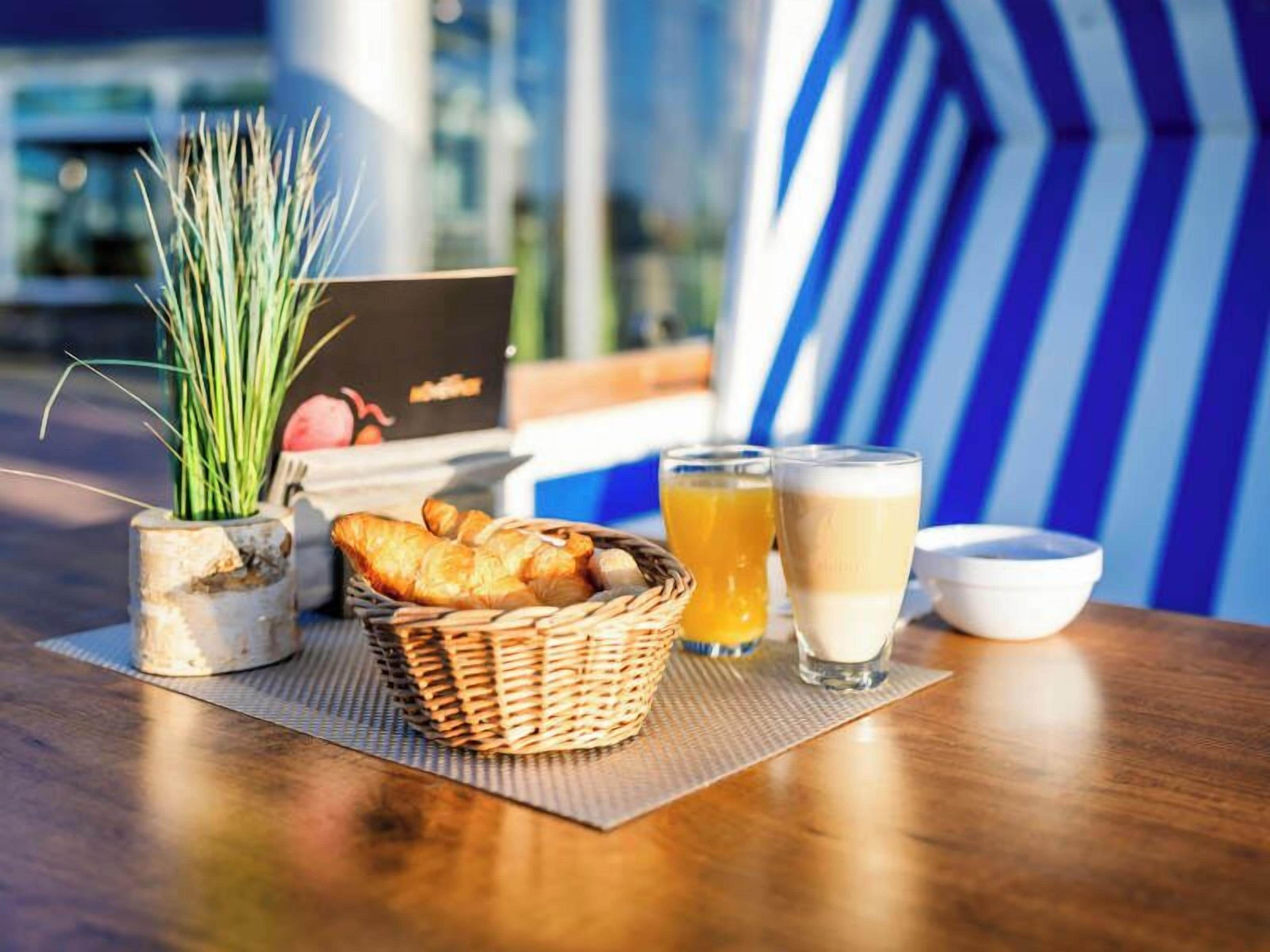 A table with a basket of croissants, a glass of orange juice, a glass of milk, and a small bowl, set on a mat with a plant and a menu in the background.