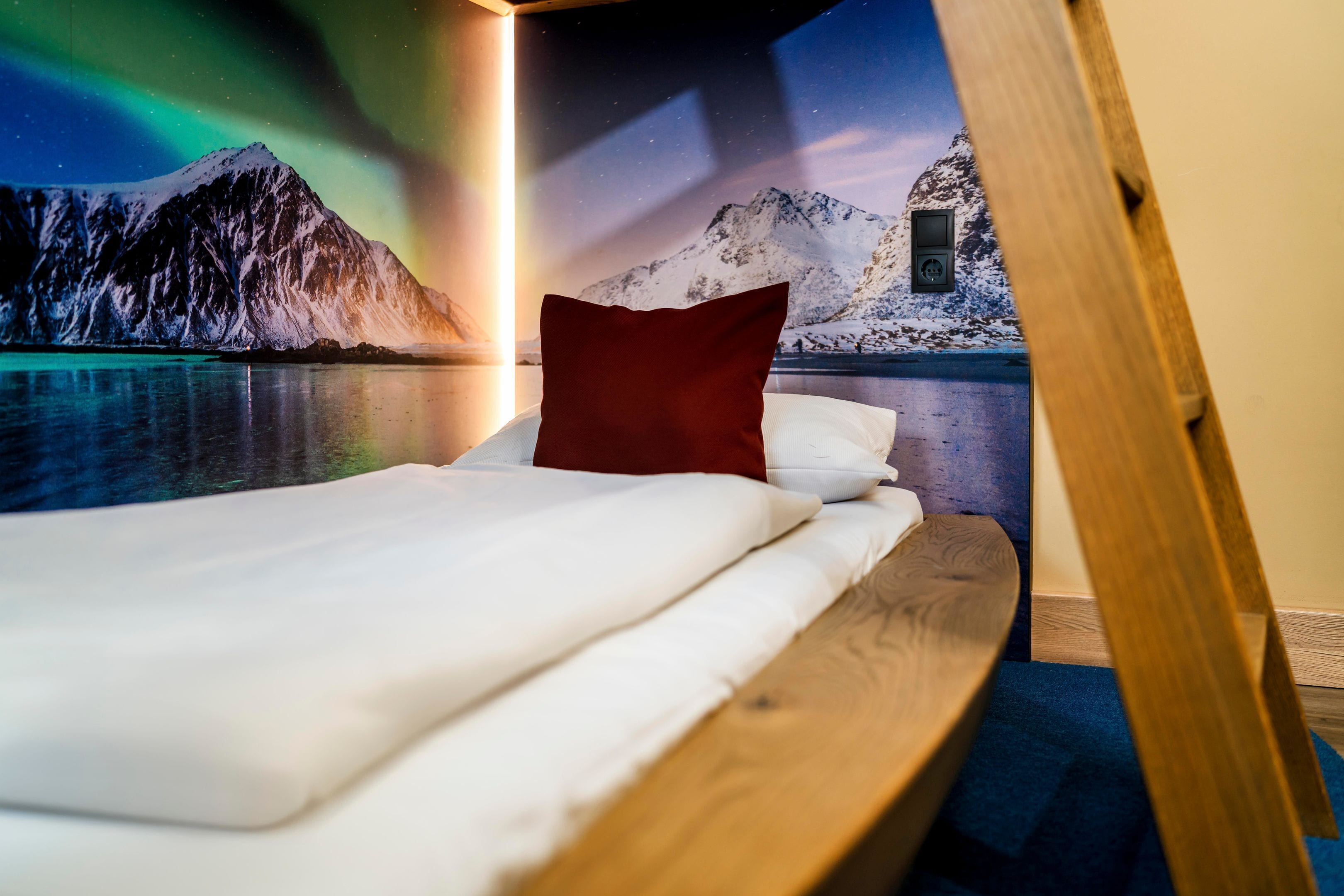 A bed with white bedding and a red pillow, set against a wall mural of a snowy mountain and lake at night.