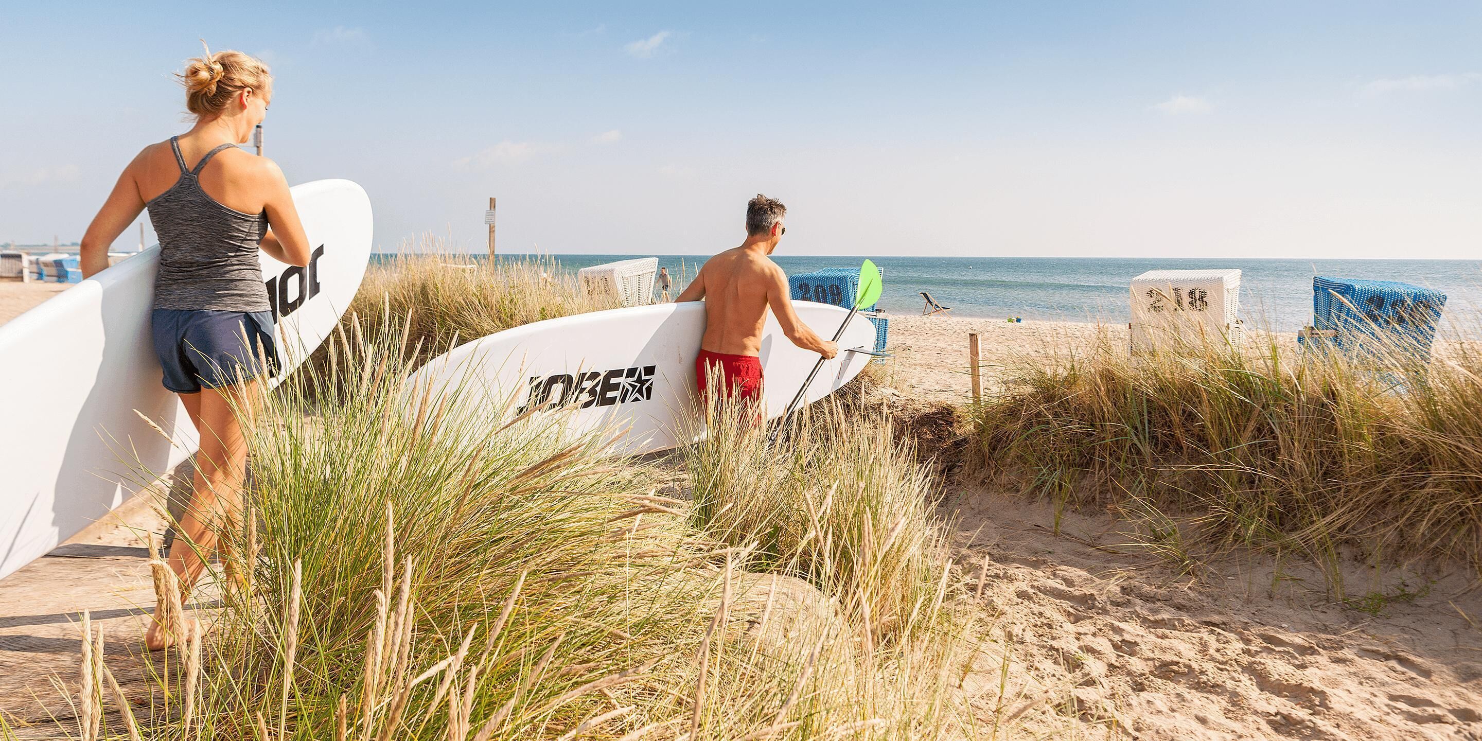 A beach with two people carrying surfboards, a sandy path, and beach chairs.