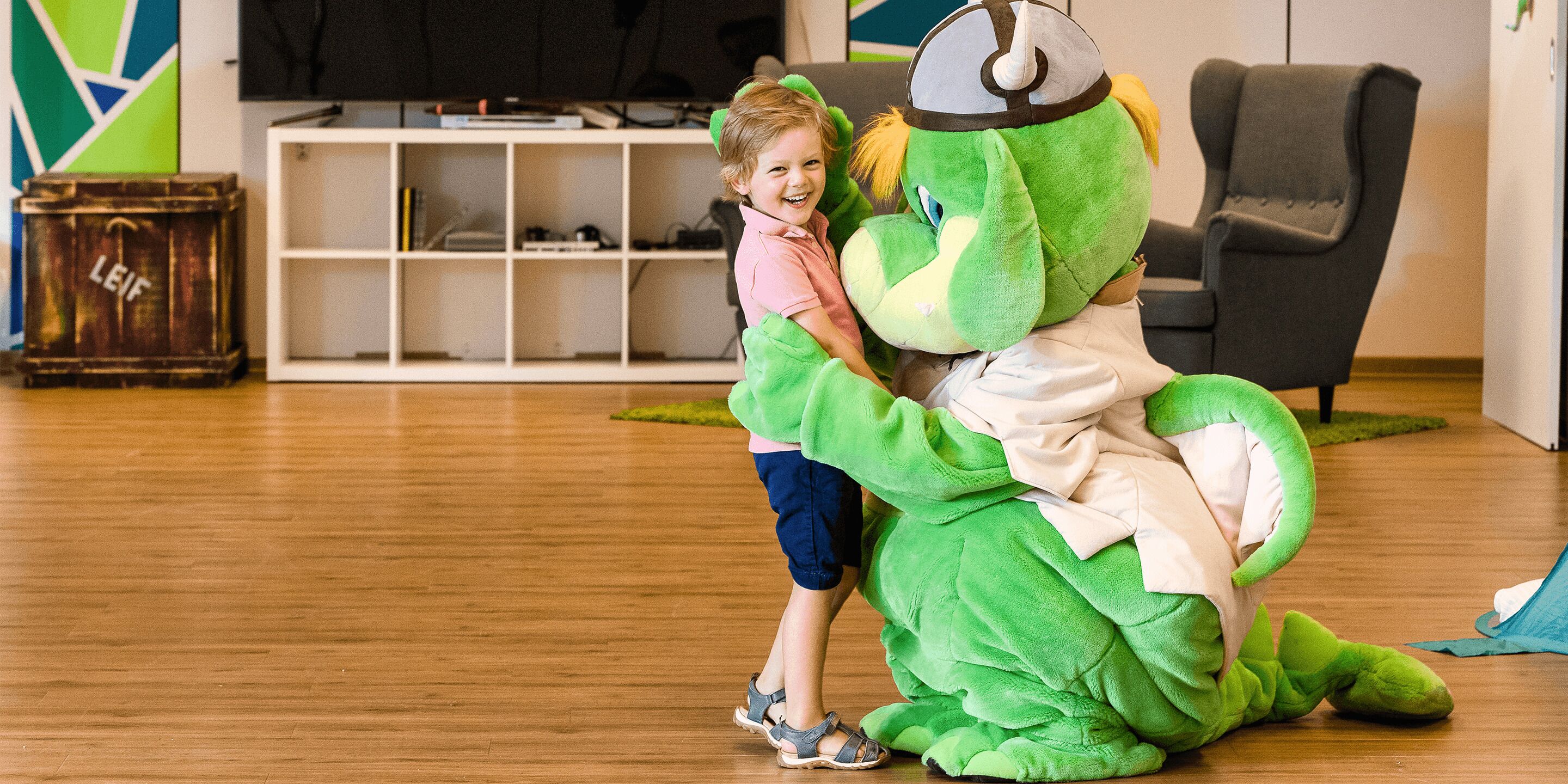 Un niño abrazando a un disfraz de dinosaurio verde en una habitación con piso de madera, un sillón gris y un mueble blanco para entretenimiento.
