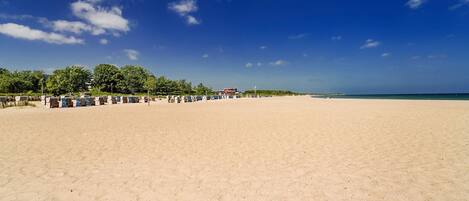 Une vaste plage de sable fin, sous un ciel bleu azur parsemé de quelques nuages. On y trouve plusieurs parasols et des chaises longues le long du rivage.