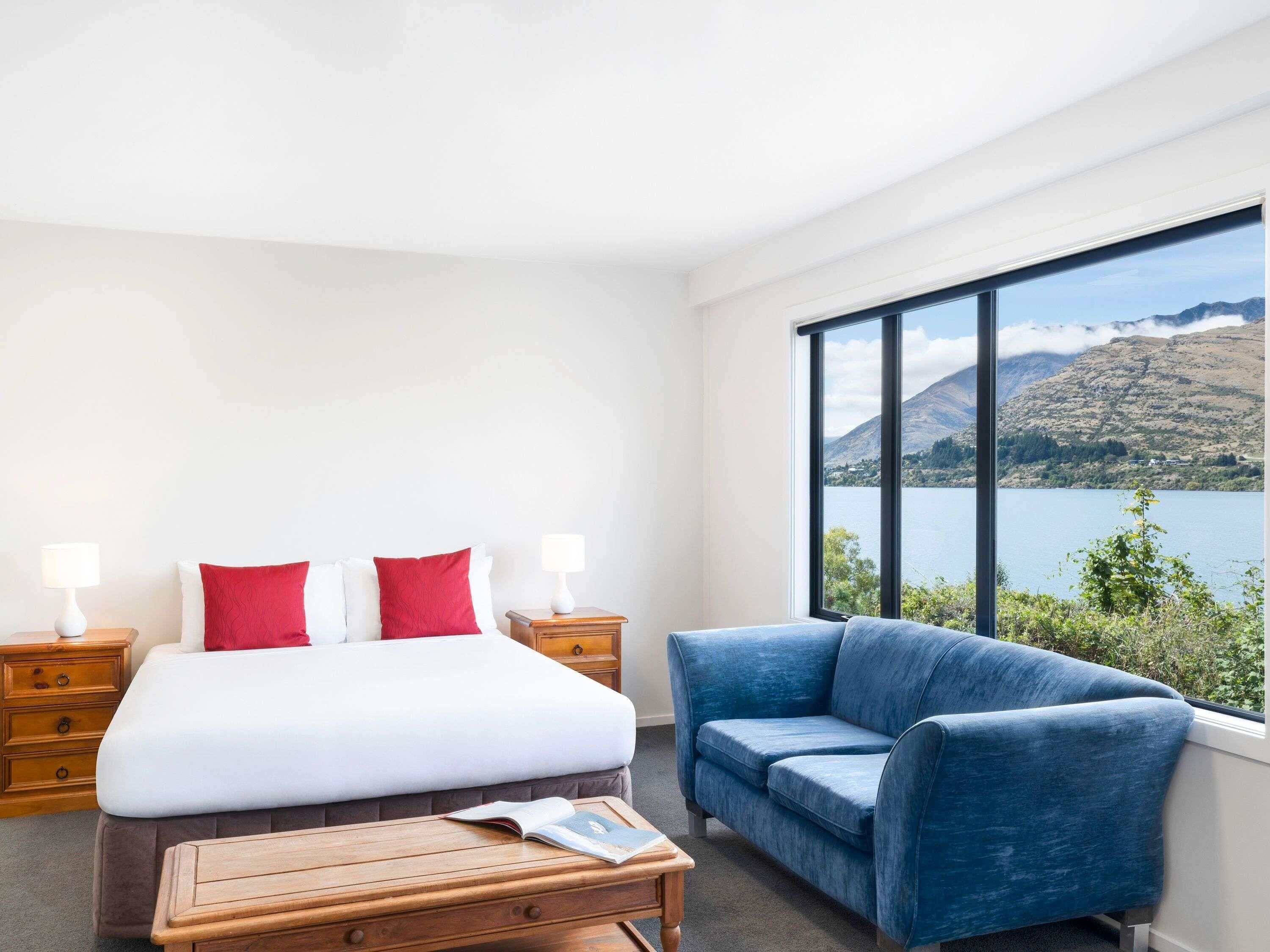 A hotel room with a bed, two bedside tables, a blue armchair, and a large window with a mountain view.