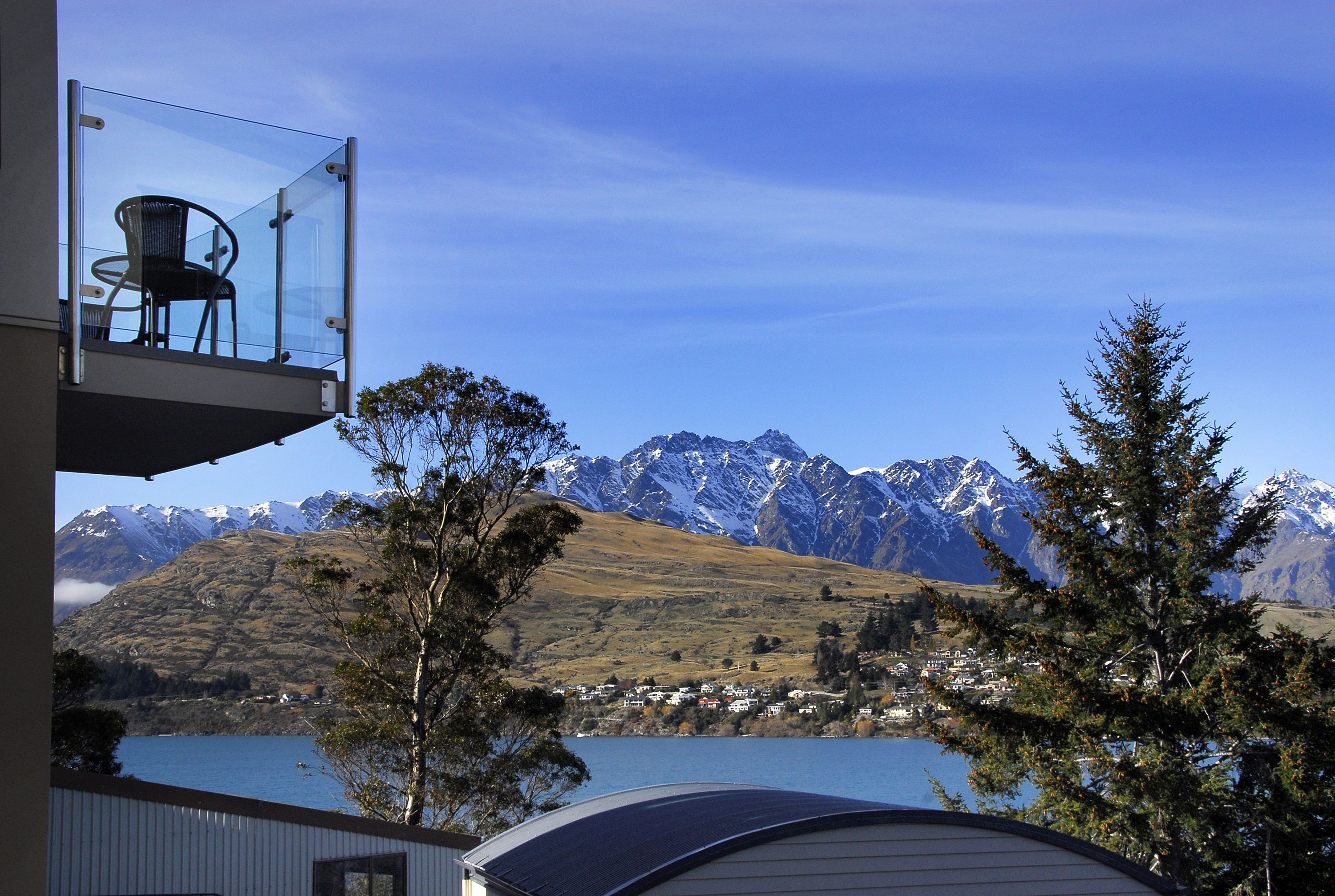 Un balcon en verre avec une chaise, offrant une vue sur un lac et des montagnes enneigées.