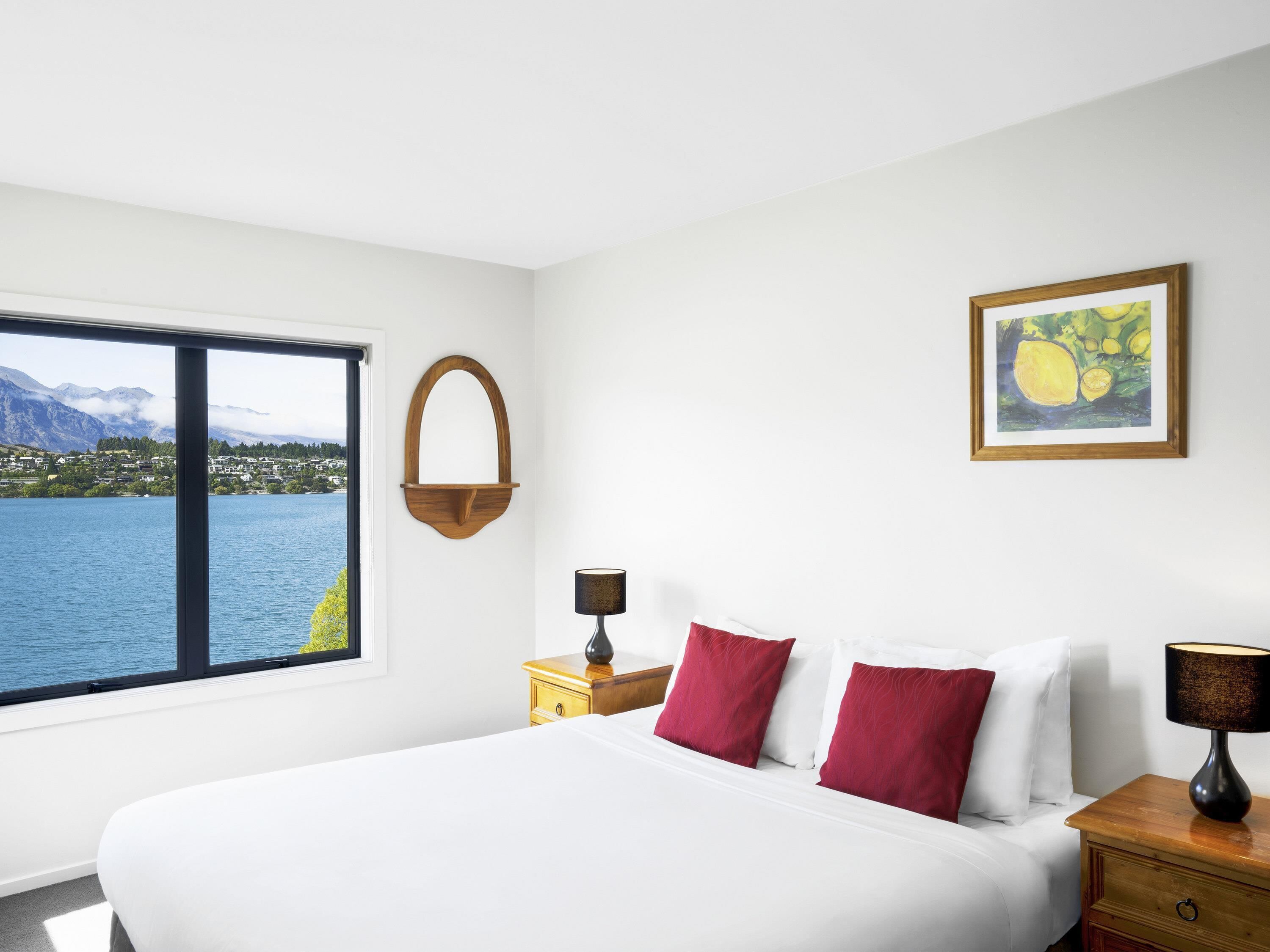 A bedroom with a bed, a nightstand, a lamp, a wall-mounted shelf, and a framed picture.