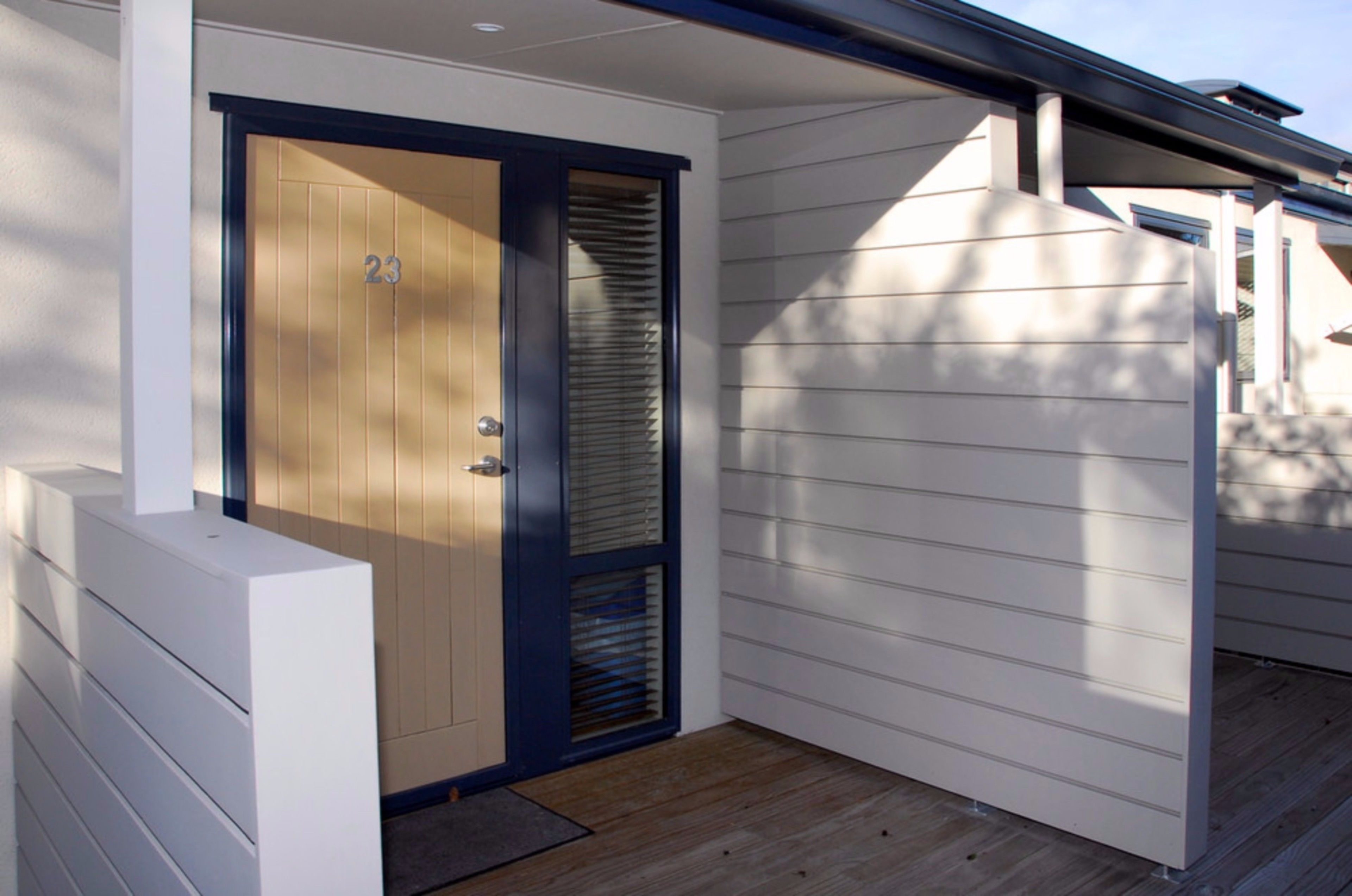 Ein modernes Haus mit einer Holztür, die mit der Nummer 23 beschriftet ist, einer Veranda mit Holzboden und weißem Außenputz.