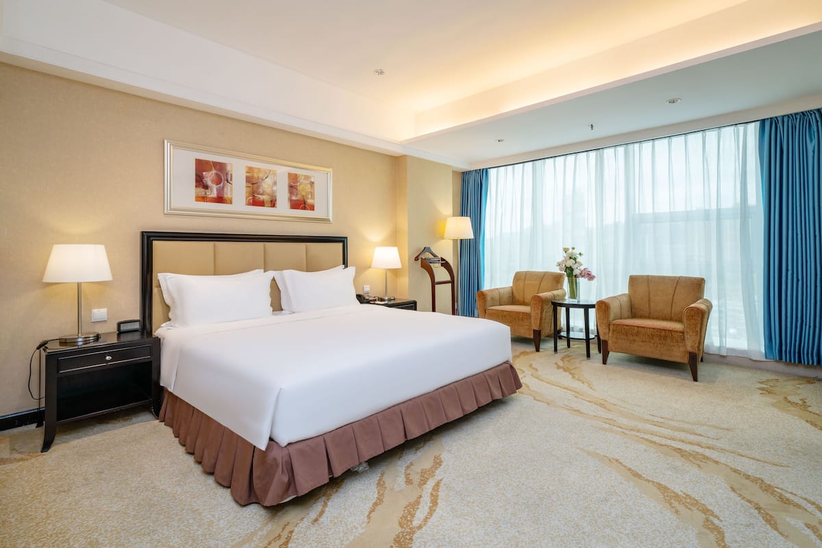 A hotel room with a large bed, two bedside tables with lamps, a chair, a small table with a vase, and a large window with curtains.