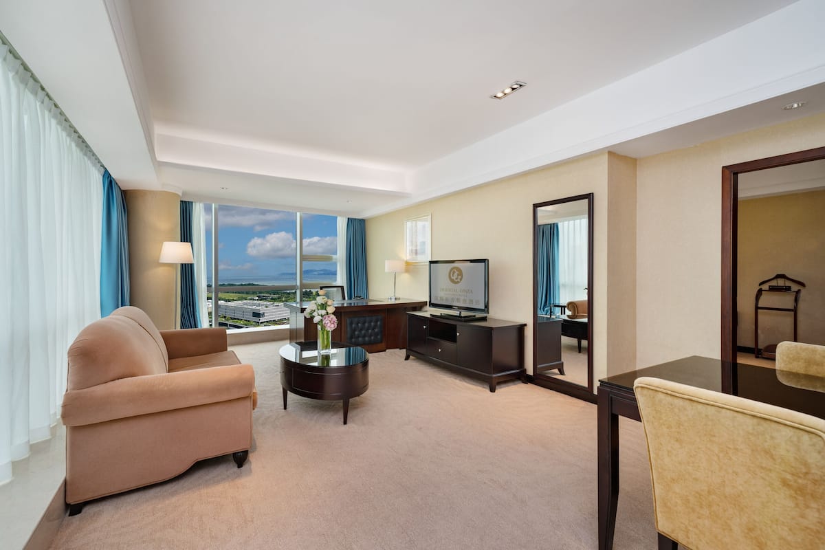 A hotel room with a large window, a sofa, a coffee table, a TV, and a mirror.