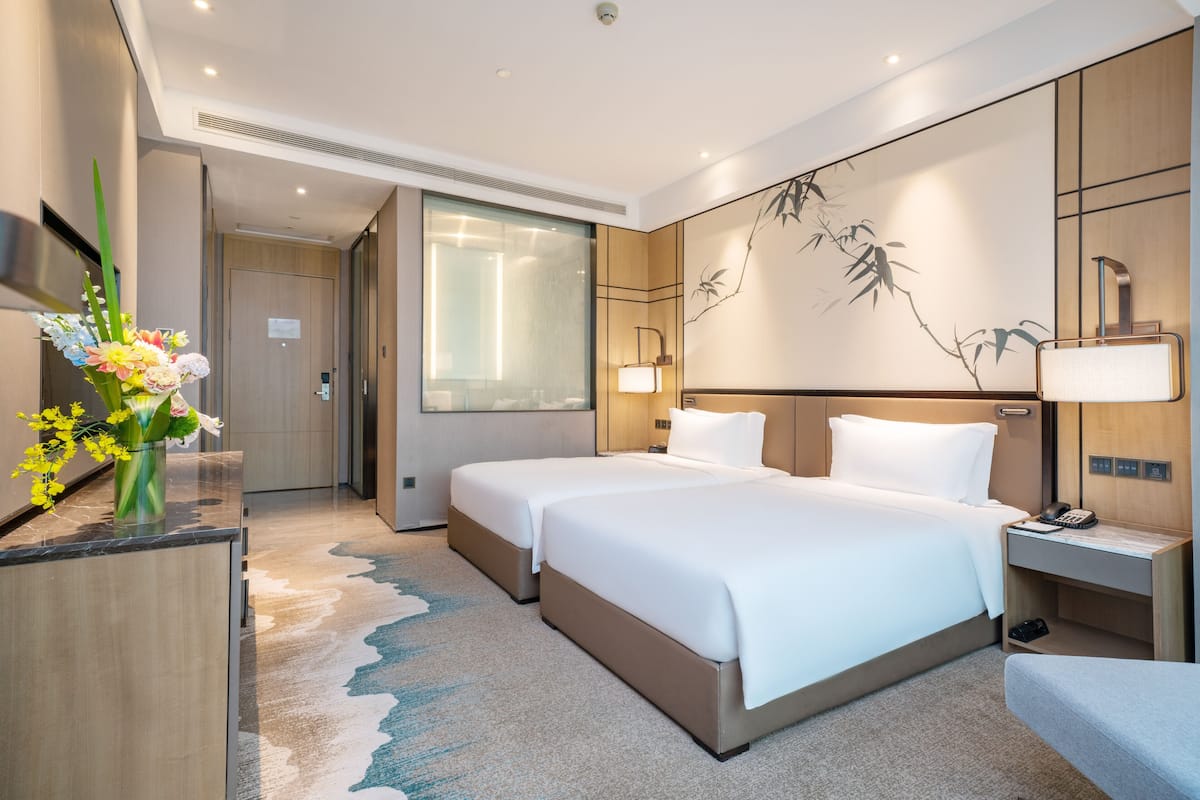 A hotel room with two beds, a desk, a chair, a vase of flowers, and a wall-mounted lamp.