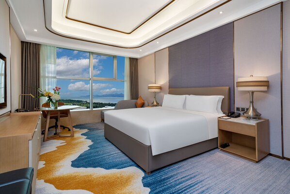Relax in a spacious room with a large bed, two bedside tables, and a desk with a chair, offering a view of the sky and distant landscape.