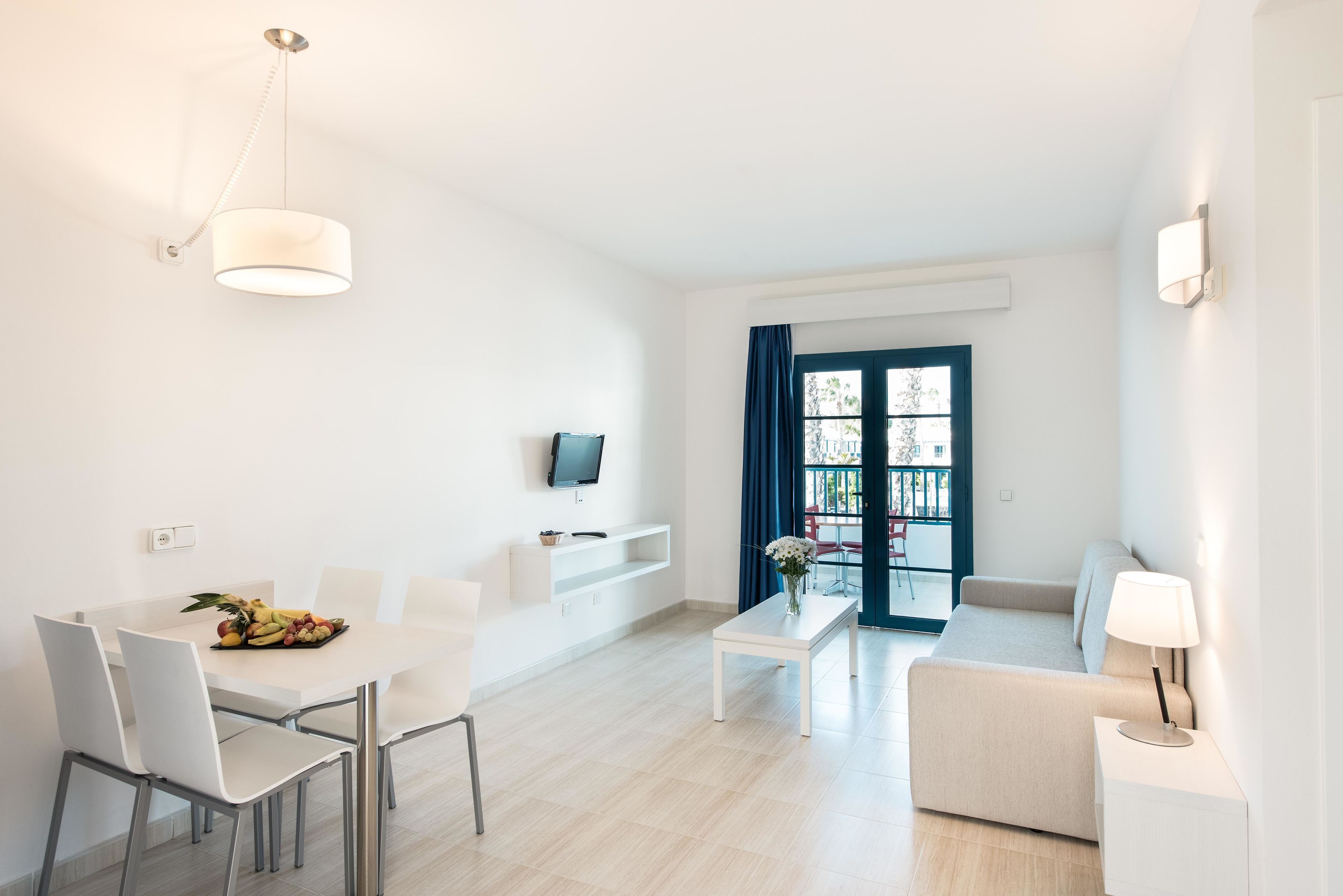 A modern living room with a sofa, a dining table with chairs, a TV mounted on the wall, and a window with curtains.