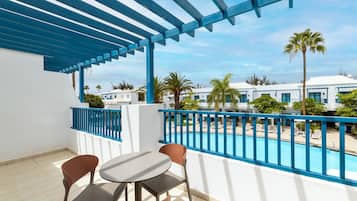 Un balcón con vista a la piscina, paredes blancas, barandillas azules y una mesa con sillas.