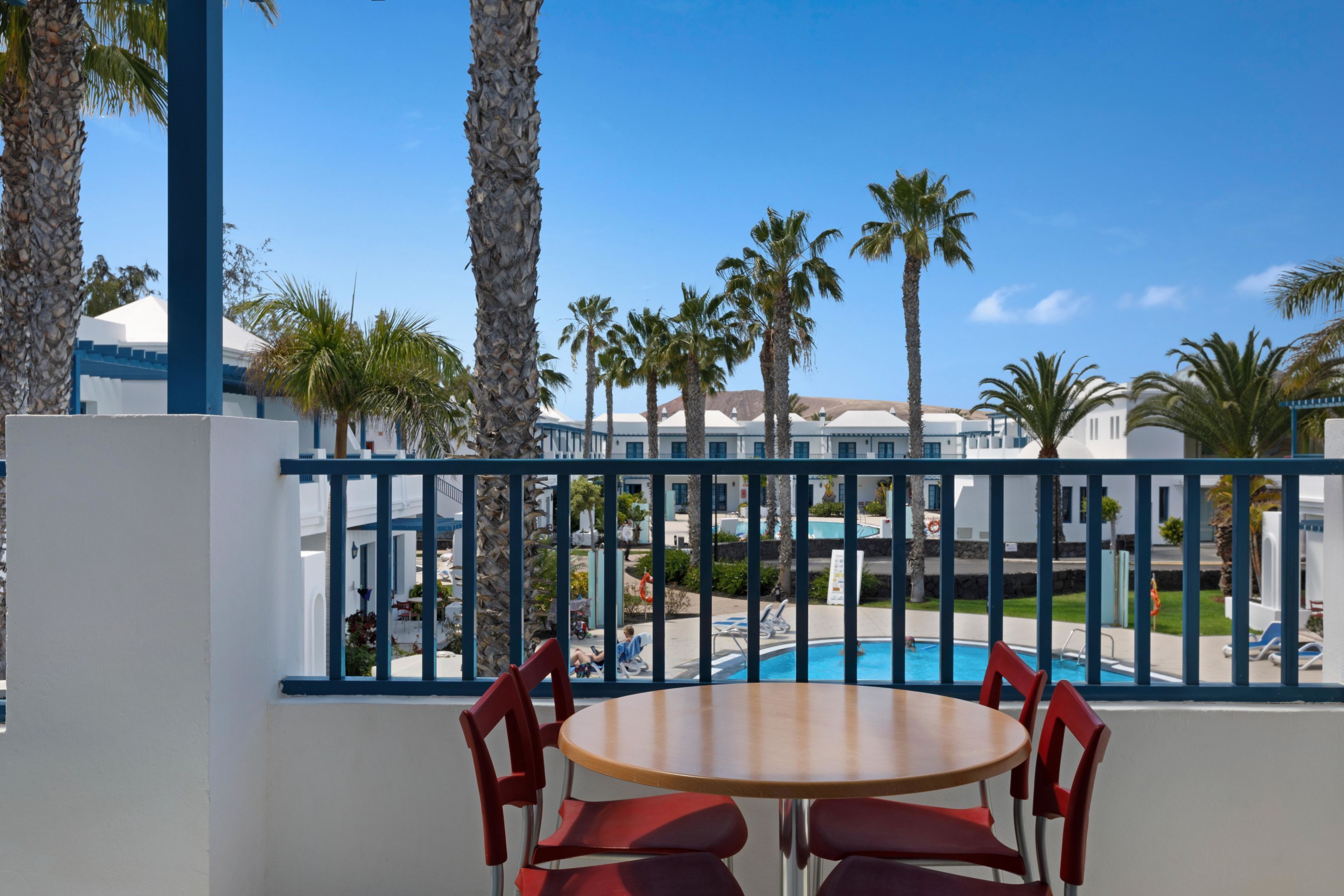 Un balcón con mesa de madera y sillas rojas, con vista a una piscina y palmeras, con edificios al fondo.