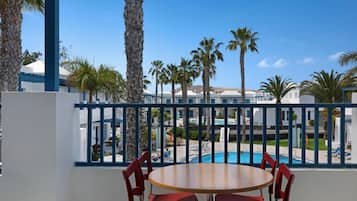 Un balcón con mesa de madera y sillas rojas, con vista a una piscina y palmeras, con edificios al fondo.