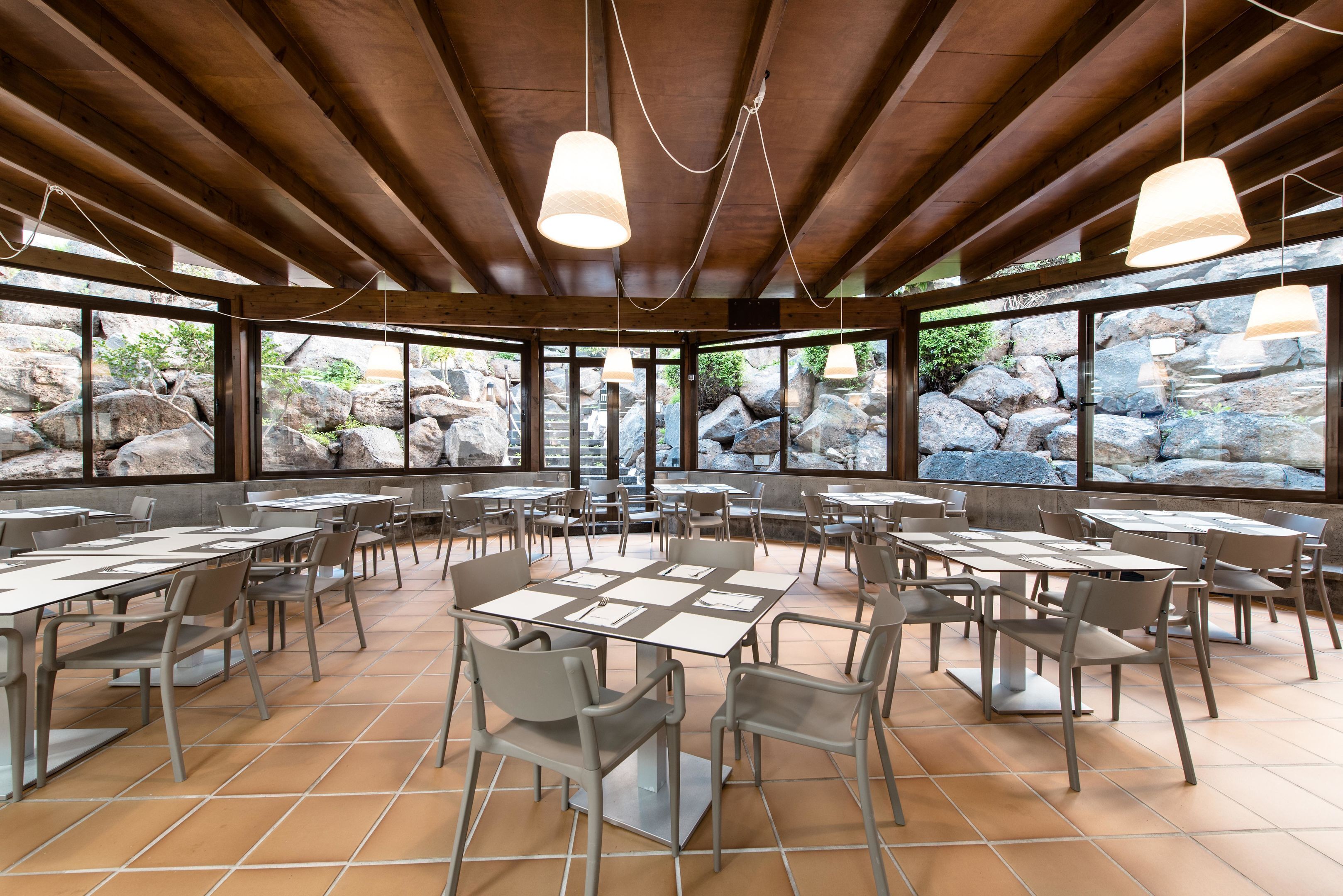 Un comedor interior con vigas de madera, grandes ventanales con vistas a las rocas y numerosas mesas y sillas.
