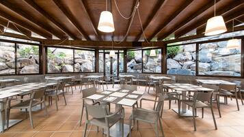 Un comedor interior con vigas de madera, grandes ventanales con vistas a las rocas y numerosas mesas y sillas.