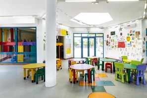 Ein helles Klassenzimmer mit mehreren Tischen und Stühlen, einem großen Fenster und Lernplakaten an den Wänden.