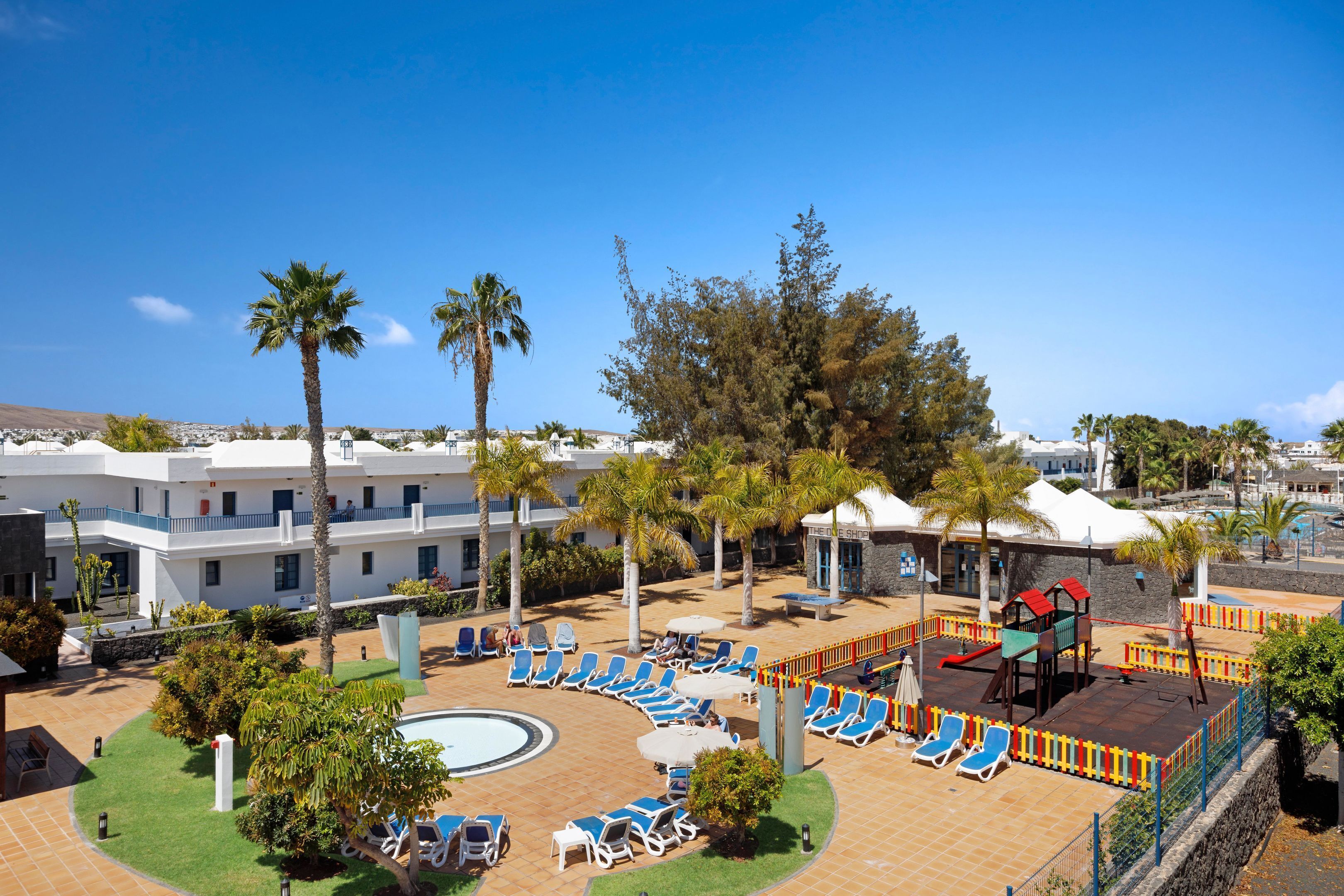 Un resort con zona de piscina, tumbonas, palmeras y parque infantil.