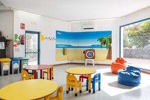 Ein hell erleuchteter Raum mit bunten Tischen und Stühlen, einem Wandgemälde mit Strandmotiv, Müllbehältern und einer Zielscheibe an der Wand.