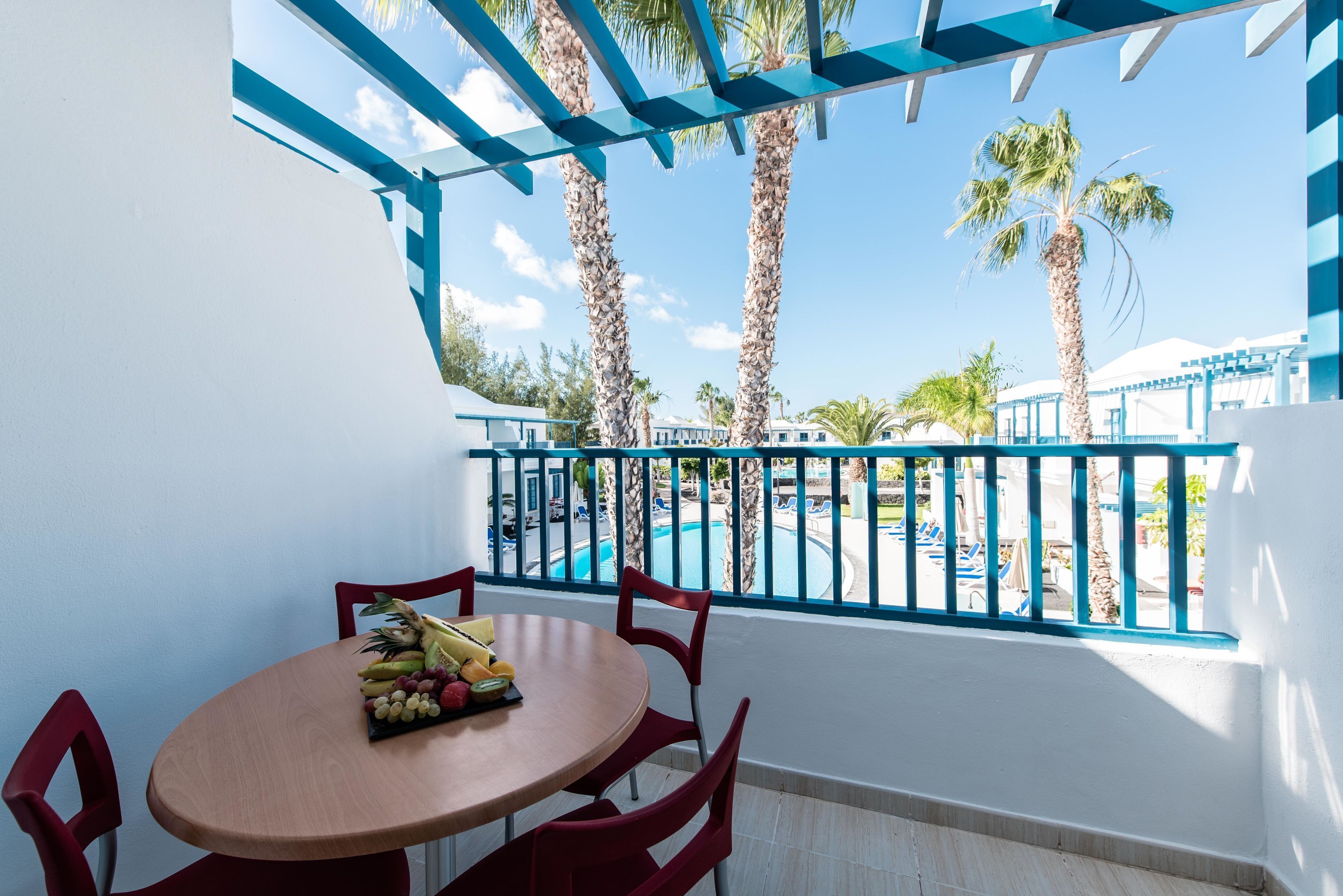 A balcony with a round table and chairs, a fruit bowl, and a view of a pool and palm trees.
