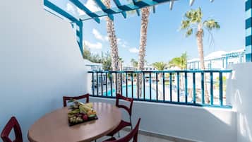 A balcony with a round table and chairs, a fruit bowl, and a view of a pool and palm trees.