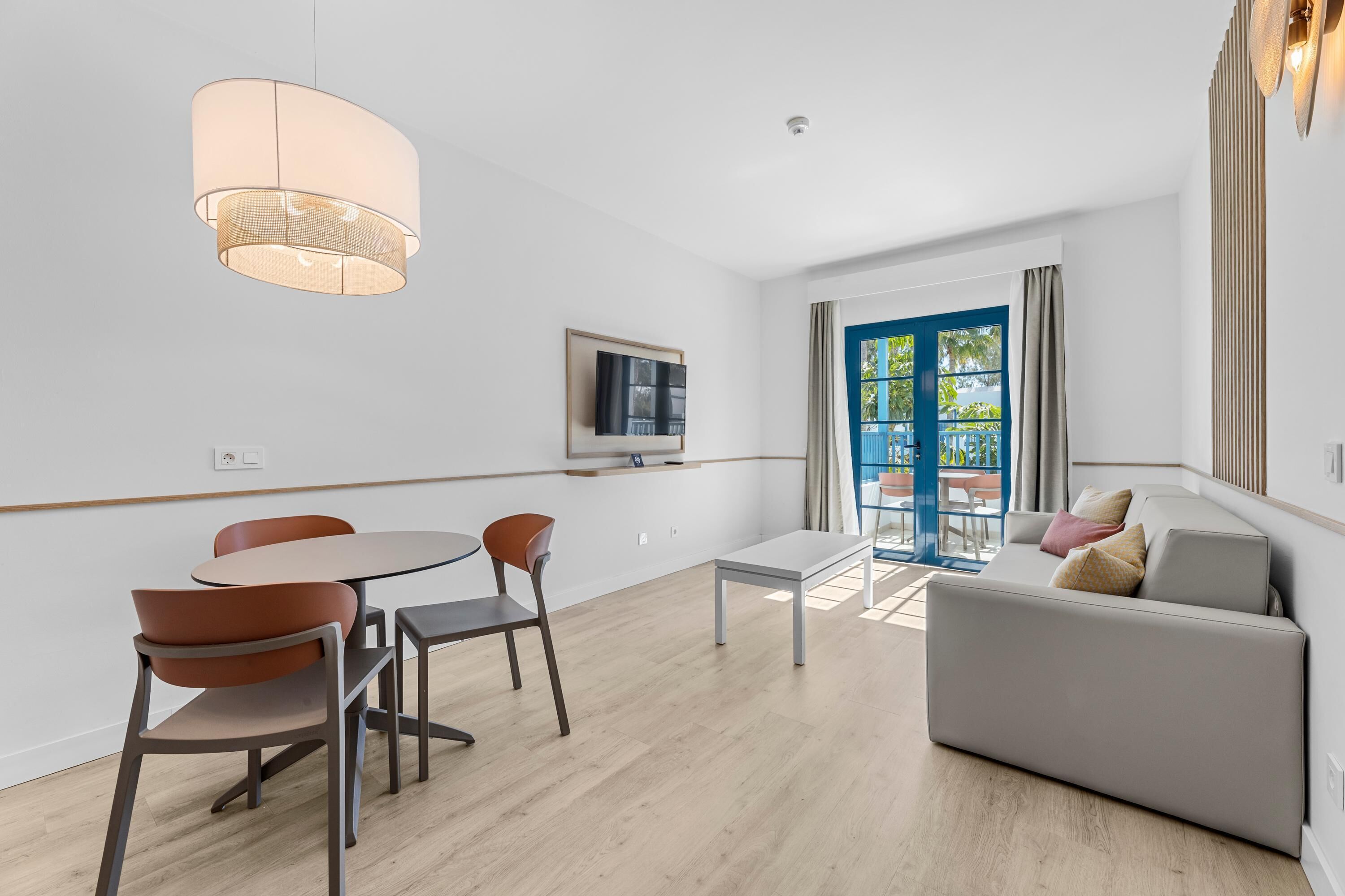 Una sala de estar moderna con un sofá, una mesa de comedor con sillas, un televisor montado en la pared y un ventanal con vistas a zonas verdes.