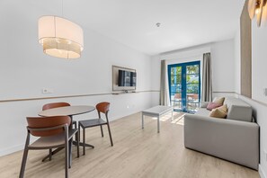 A modern living room with a sofa, a dining table with chairs, a TV mounted on the wall, and a large window with a view of greenery. - THB Tropical Island Aparthotel (Yaiza)