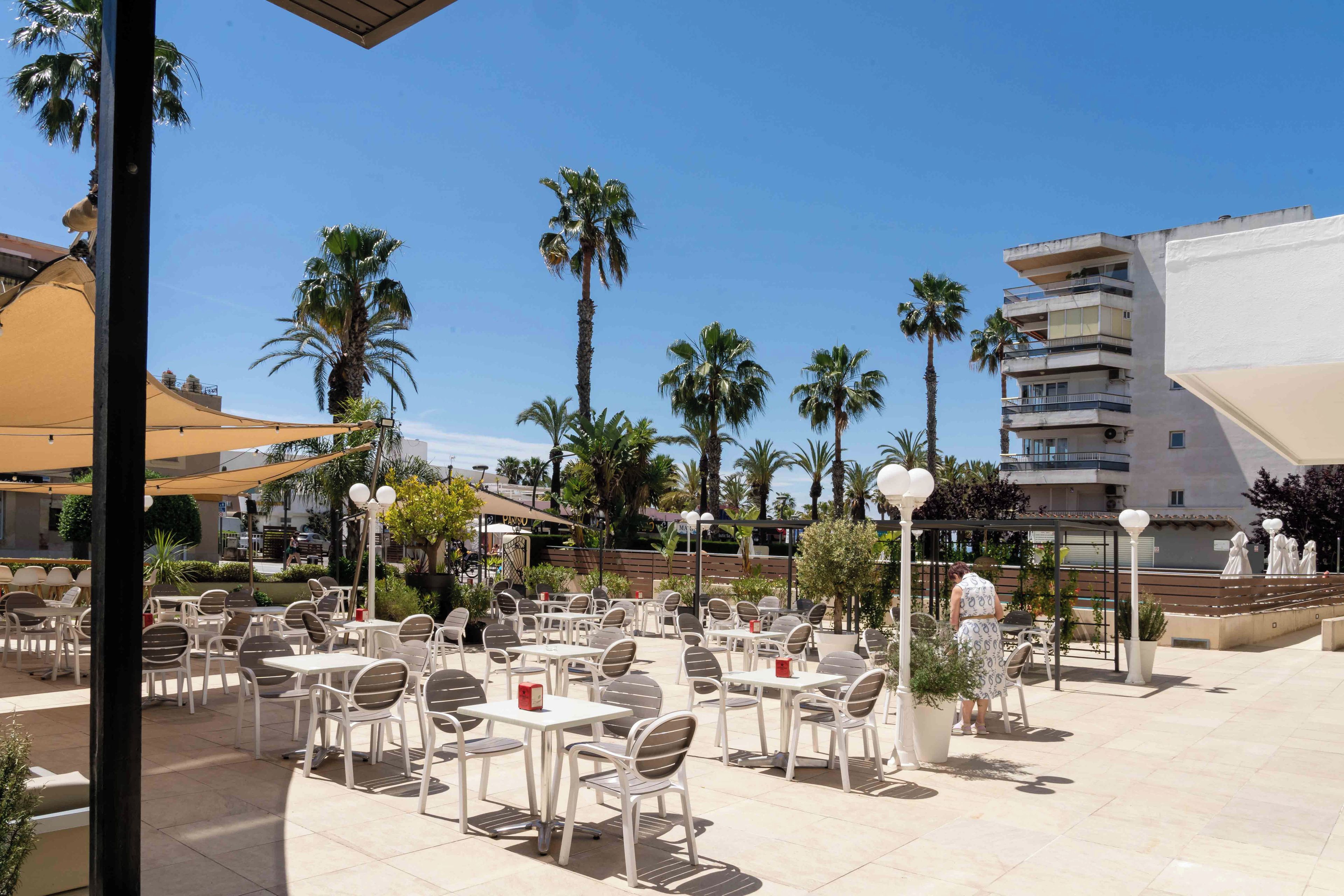 An outdoor patio area with white tables and chairs, palm trees, and a building in the background.