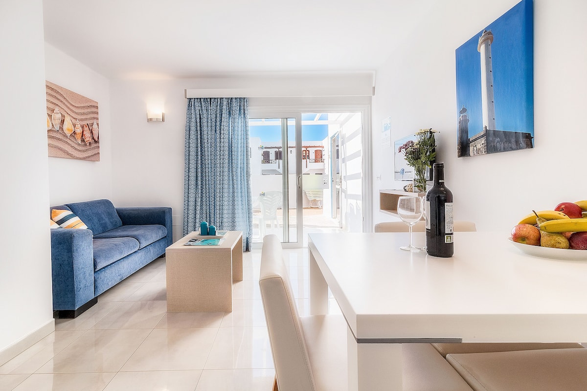 Una sala moderna con un sofá azul, una mesa de centro, un comedor con una mesa blanca y vistas a través de la puerta de cristal.