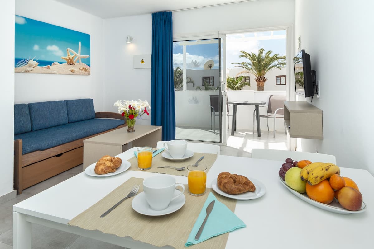 Habitación de hotel moderna con sofá, mesa de desayuno y balcón con vista a palmeras.