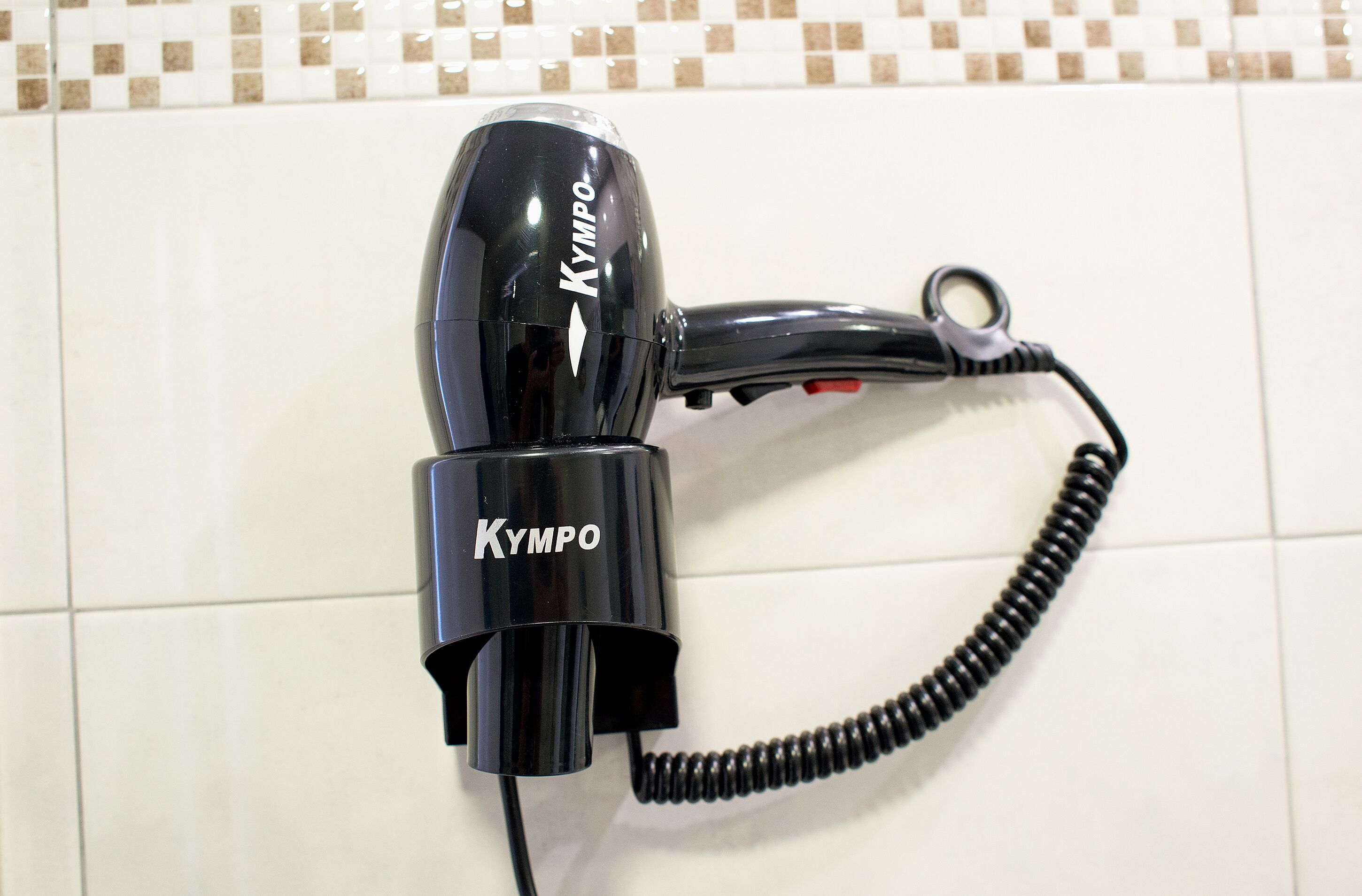 A black hairdryer with a coiled cord, labeled "Kympo," on a tiled floor.