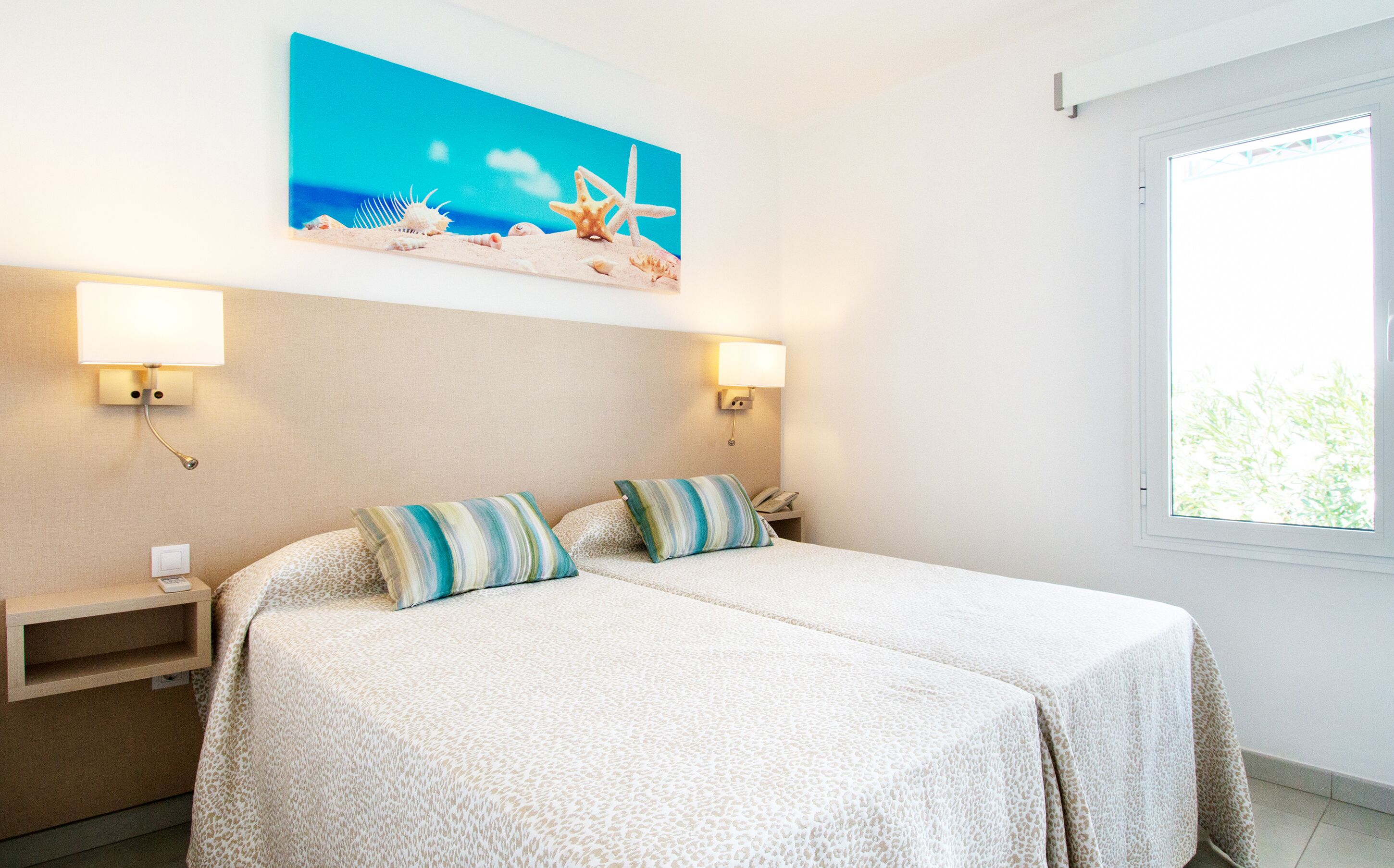 A hotel room with a bed, two pillows, a nightstand, a lamp, a painting of a beach scene, and a window.
