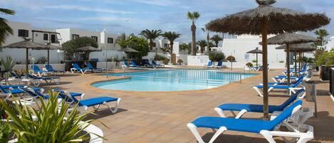 A pool area with sun loungers and umbrellas, surrounded by palm trees and white buildings.
