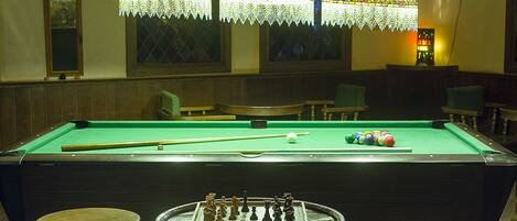 Une table de billard avec un jeu d’échecs placé au centre, entourée de tabourets, le tout sous trois lampes suspendues.