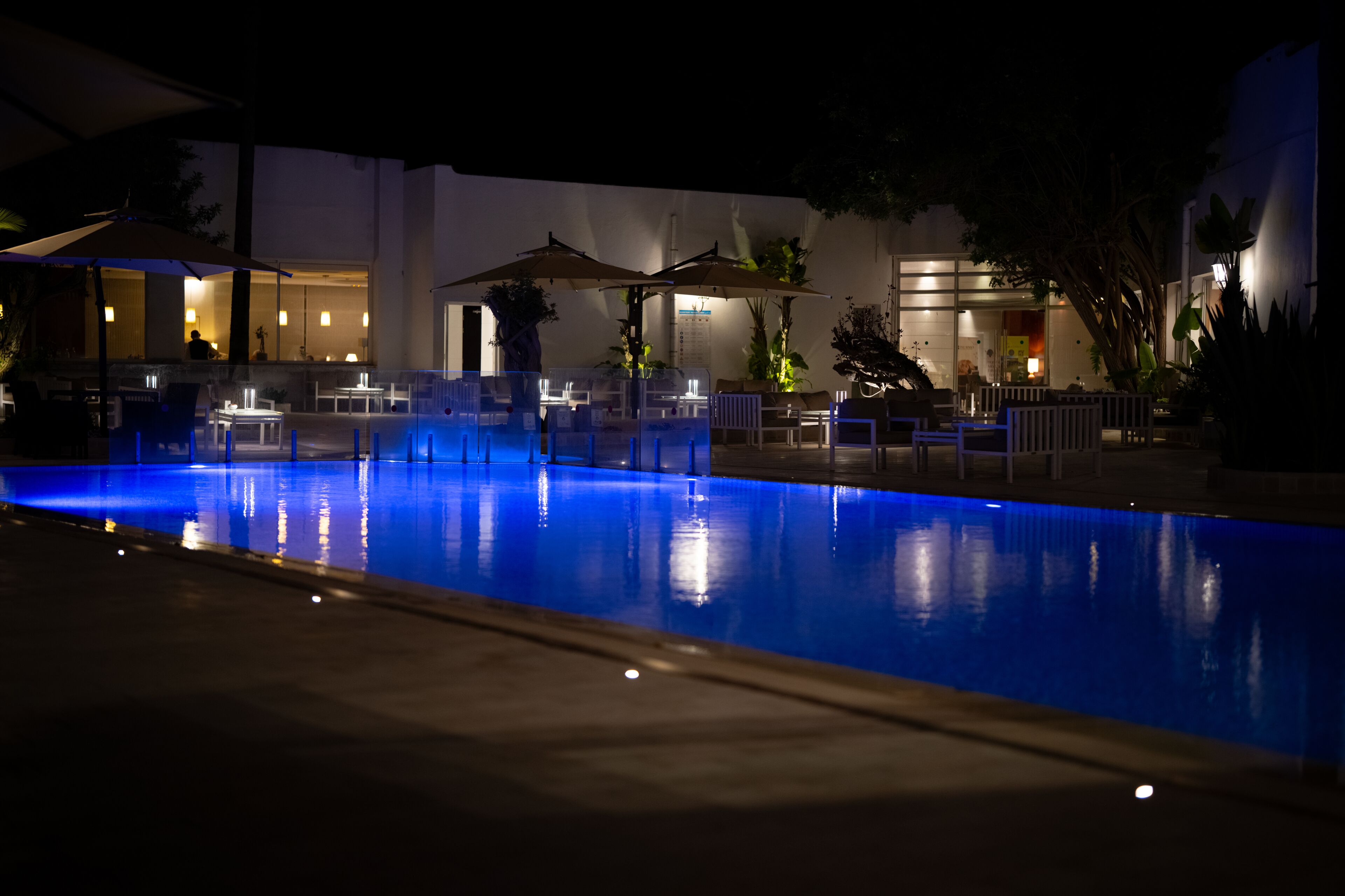 Una piscina nocturna con sillones de descanso y sombrillas, rodeada por un edificio con ventanas y puertas visibles.