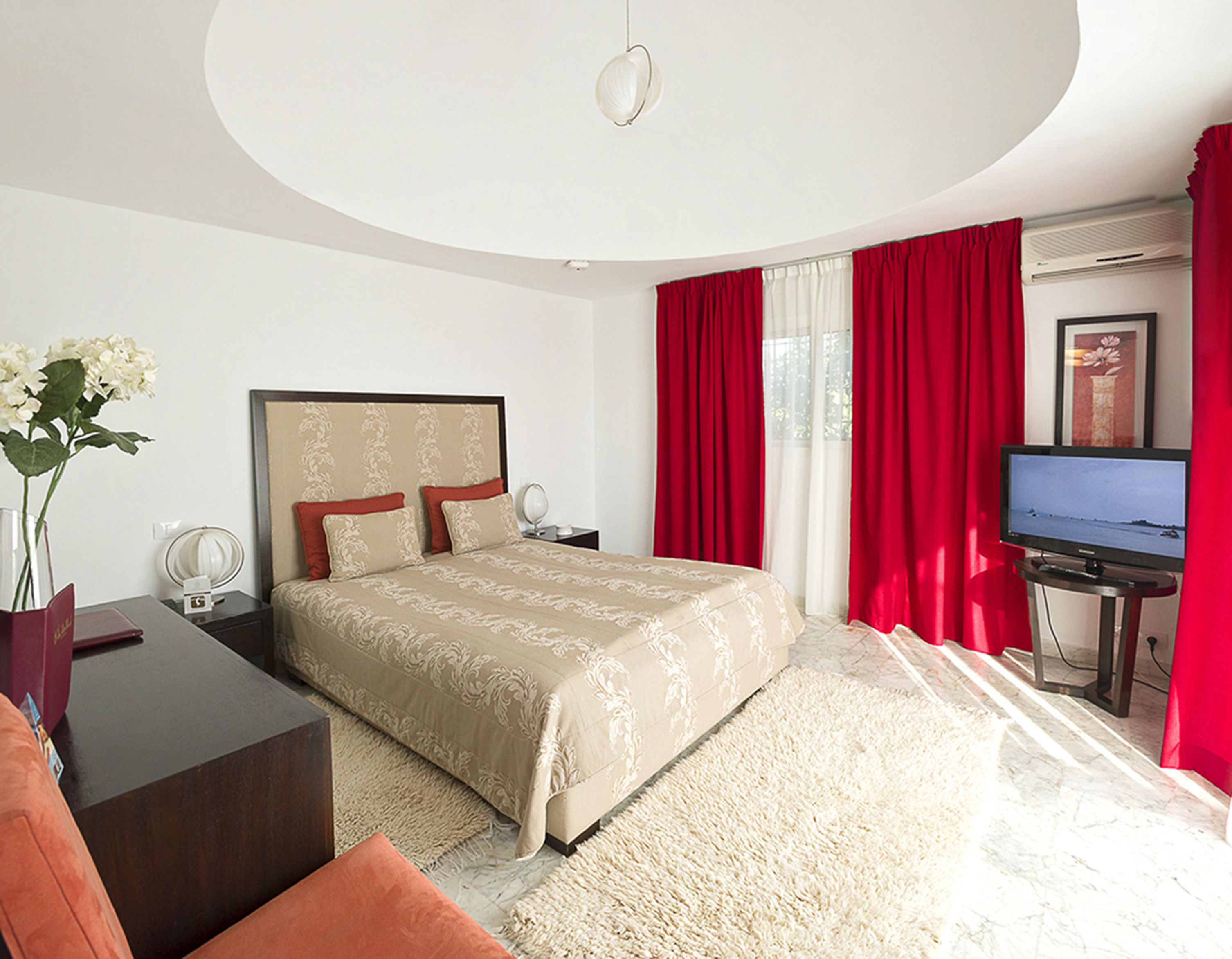 Une chambre d’hôtel avec un lit, un bureau, une télévision et une grande fenêtre avec des rideaux rouges.