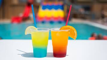 Two colorful drinks with straws and a slice of lemon on the left and an orange slice on the right, placed on a white surface with a pool and a colorful inflatable in the background.