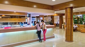 Zona de recepción de un hotel con mostrador de recepción identificado como "RECEPCIÓN", una vitrina de vidrio y un pilar grande.
